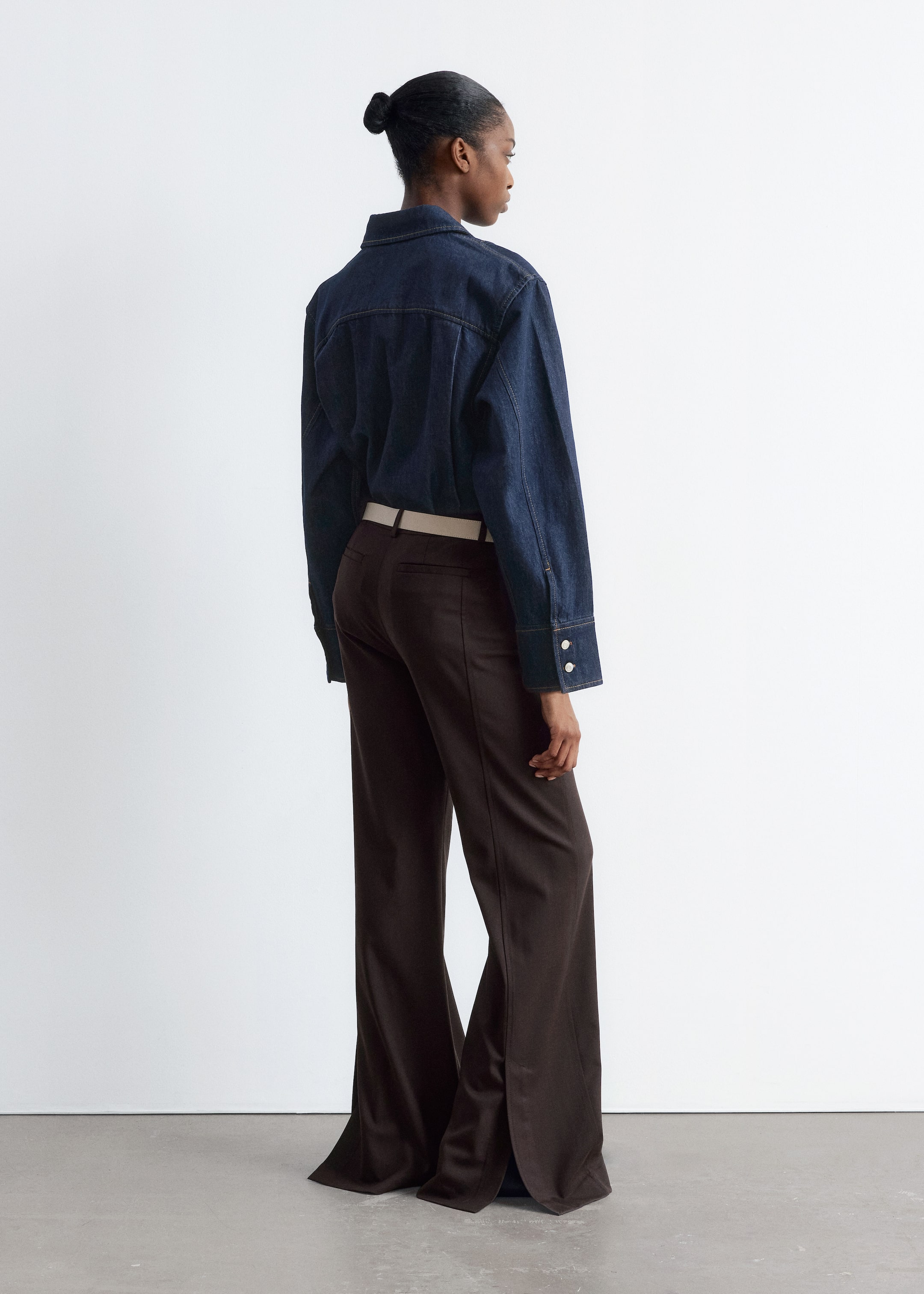 Agrandir l'image: Une chemise en denim bleu foncé, avec un col et des poignets boutonnés, est portée sur un pantalon marron foncé, taille haute et à jambes larges. Le pantalon présente une ceinture de couleur claire et des fentes distinctives sur l'ourlet latéral, l'ensemble étant présenté de dos, légèrement en angle.