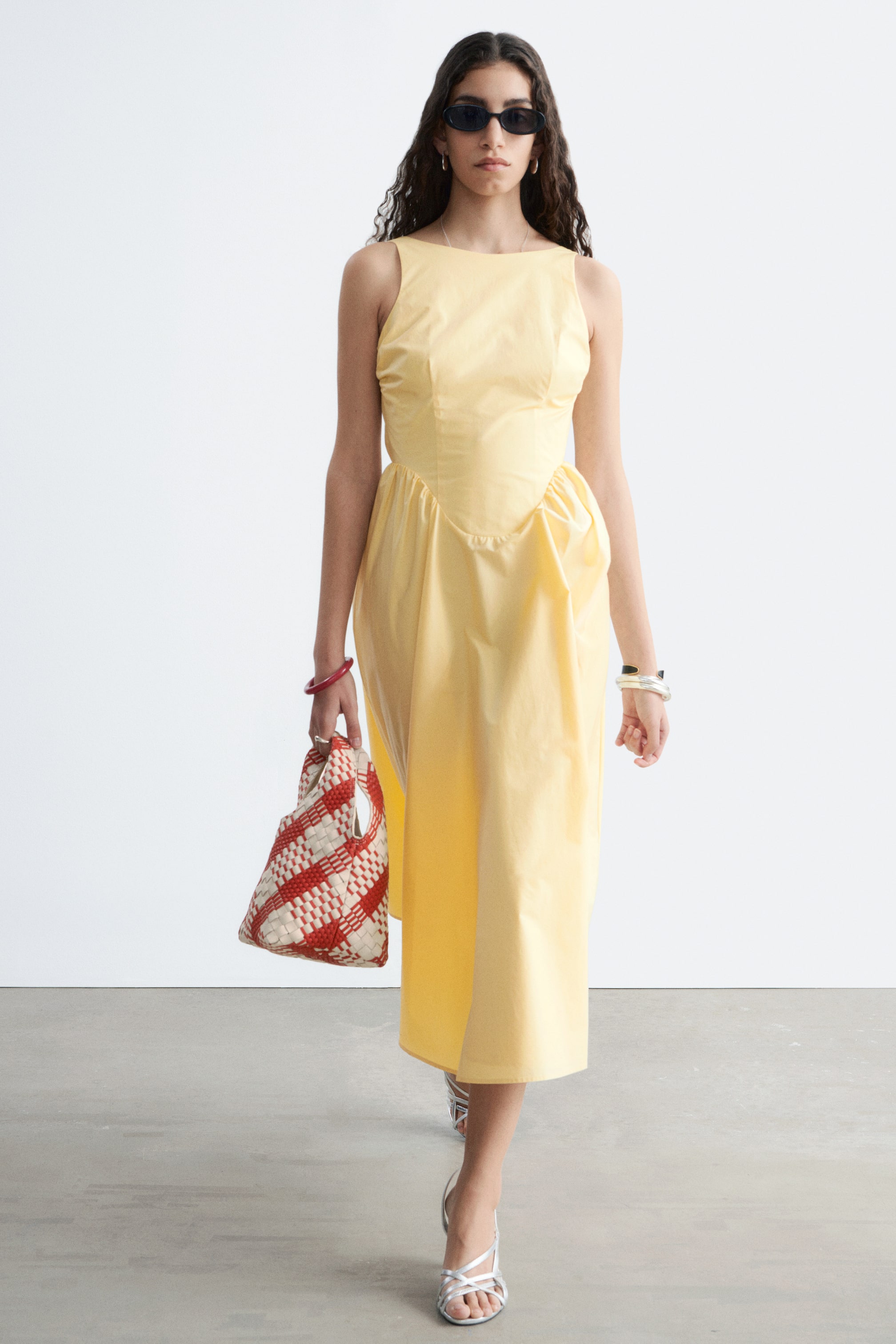 Größeres Bild anzeigen: Ein gelbes ärmelloses Midikleid mit hohem Ausschnitt und figurbetontem Oberteil. Der Rock ist an der Taille gerafft und fällt dramatisch zur Seite, wodurch eine asymmetrische Silhouette entsteht.