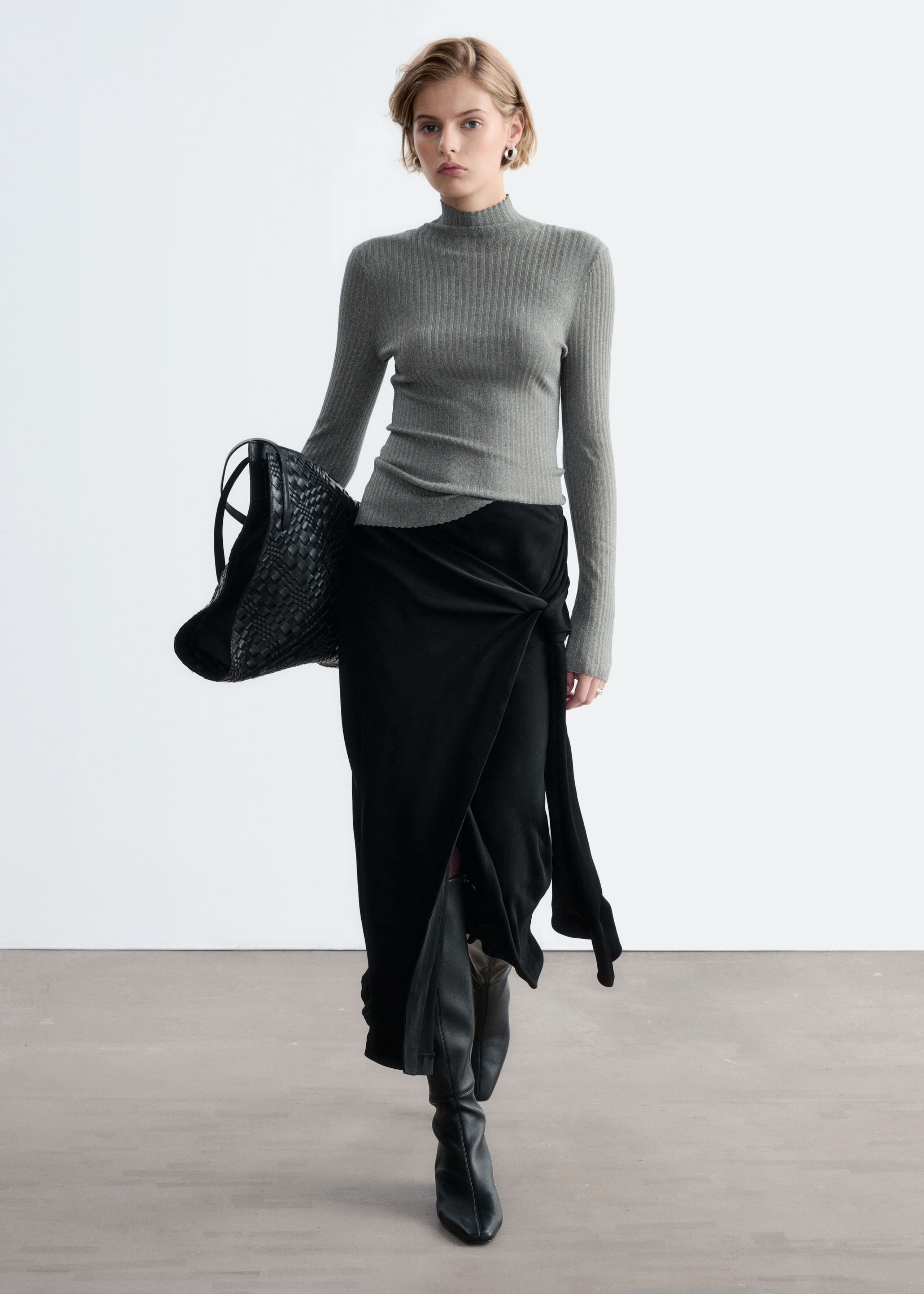 A gray, rib-knit mock neck top with a subtle metallic sheen and scalloped edges is worn by the model. The long-sleeve top is styled with a draped black wrap skirt, tall black leather boots, and a large black woven bag carried under the arm.
