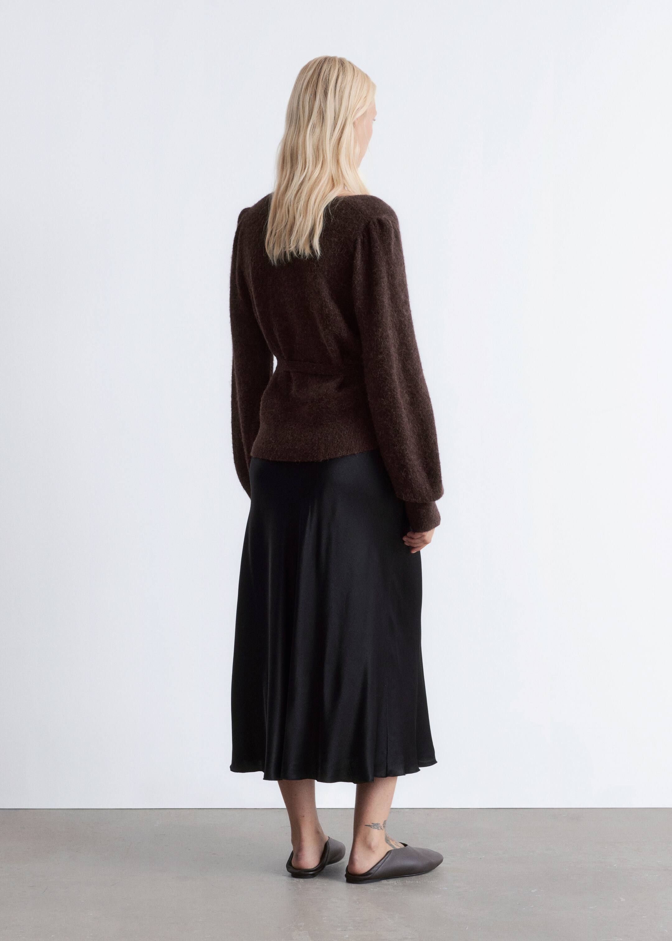 Ingrandisci l'immagine: Un top avvolgente a maglia con texture marrone scuro, presentato da dietro e leggermente angolato a destra, presenta maniche lunghe voluminose con polsini aderenti e una cintura a nastro che stringe la vita. È stratificato su una gonna midi nera fluente, che arriva a metà polpaccio, e abbinato a mule piatti grigio scuro.