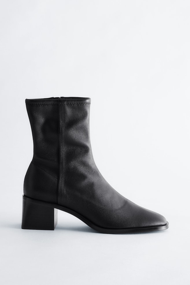 Une botte noire à la cheville présente une tige lisse et ajustée avec un bord supérieur élastiqué et une fermeture éclair latérale visible. Elle arbore un bout carré et un talon bloc noir. - 1