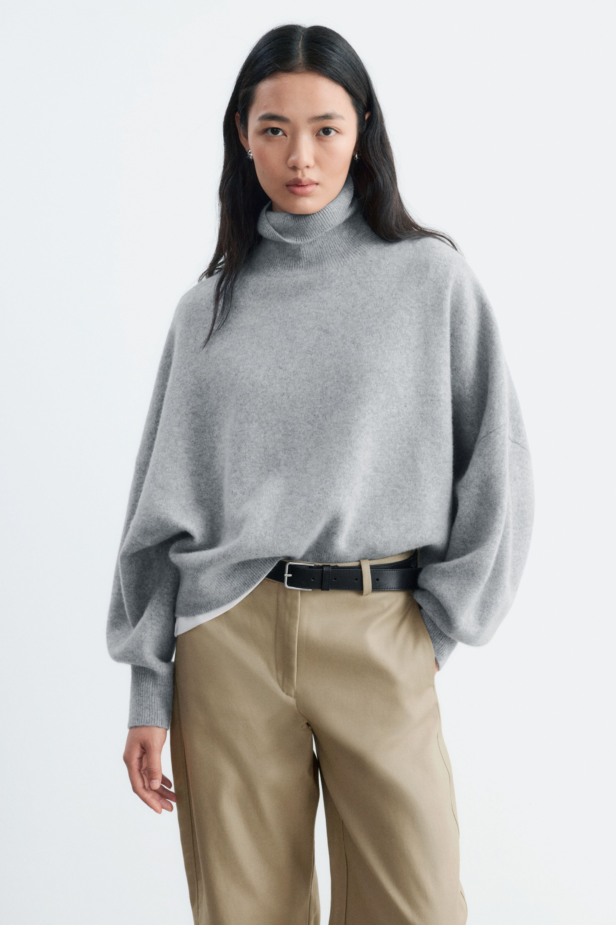 View larger image: An oversized grey knit jumper featuring a folded ribbed roll neck and dropped shoulders is being worn by the model. The jumper is styled partially tucked into beige trousers with a black belt.