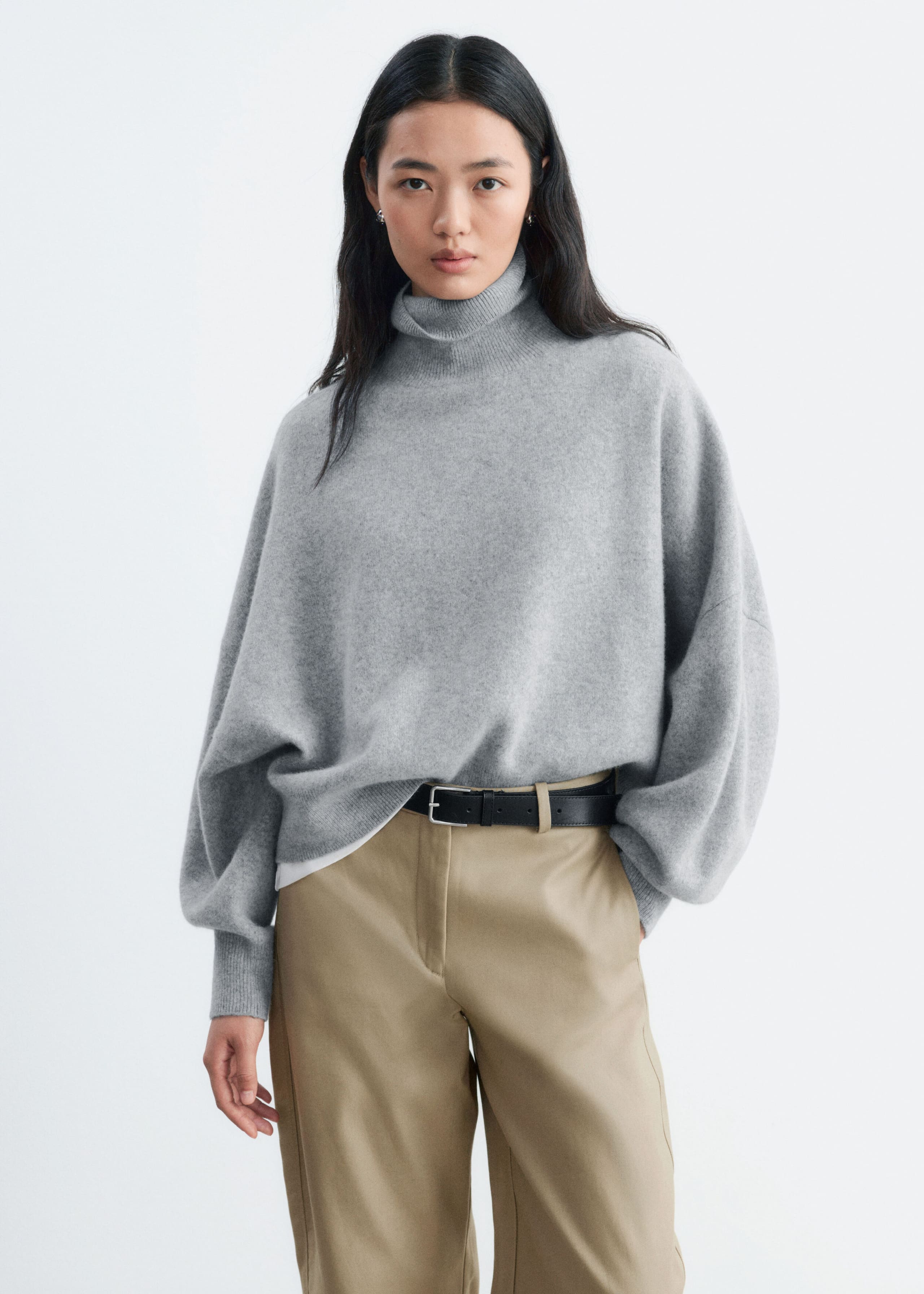 An oversized grey knit jumper featuring a folded ribbed roll neck and dropped shoulders is being worn by the model. The jumper is styled partially tucked into beige trousers with a black belt.