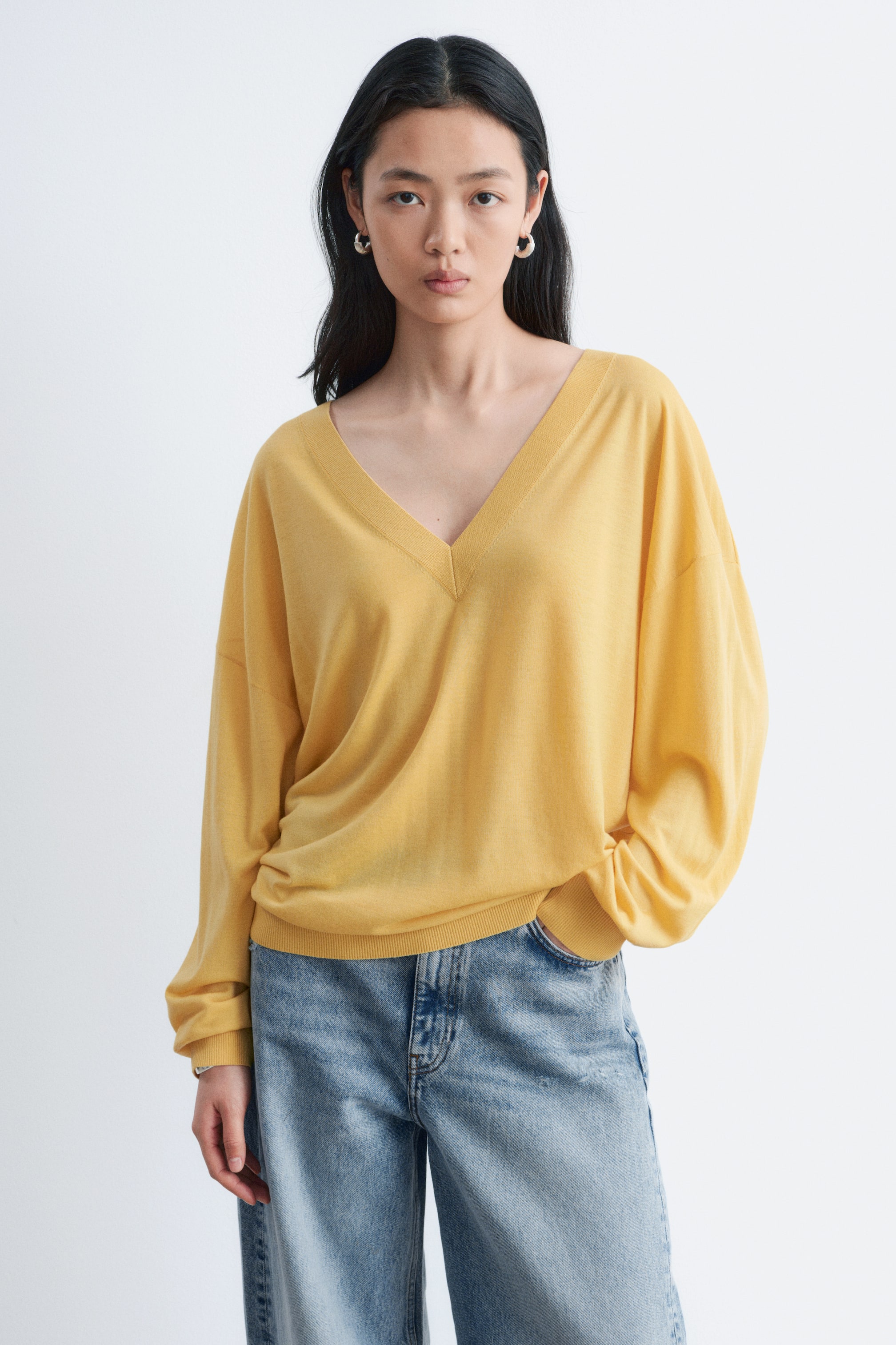 הצגת תמונה גדולה יותר: סוודר צהוב עם מחשוף V מציג גזרה רפויה עם מחשוף V עמוק. השרוולים הארוכים והנפחיים שלו מופיעים מעט מקובצים באזור האמות, ושולי הסרוגים מונחים בגובה הירך.