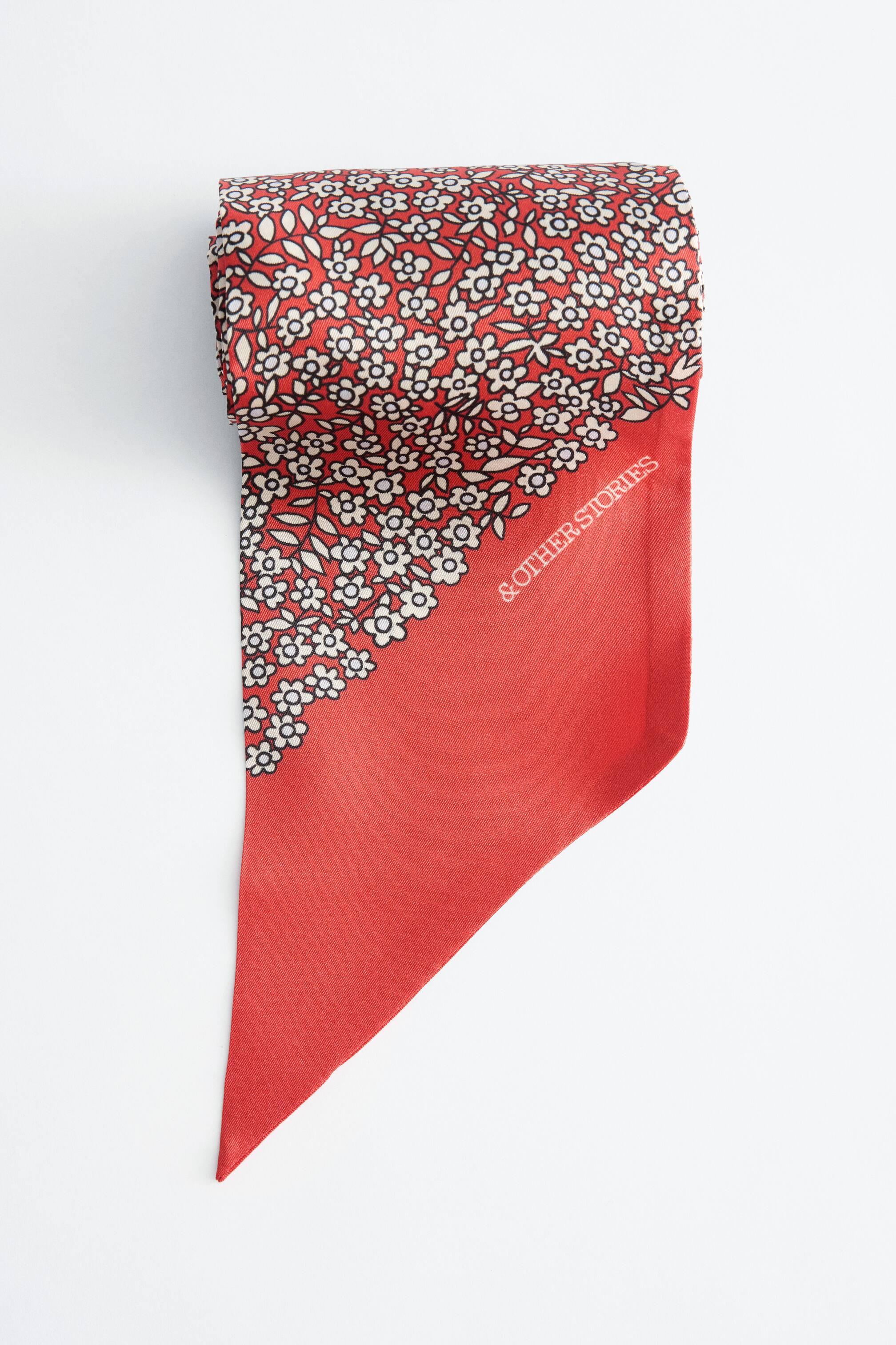 Se større billede: Et slankt tørklæde vises med den øverste del rullet, der viser et rødt stof prydet med et delikat hvidt og sort blomster mønster. Mønsteret danner en diagonal kant med en ensfarvet rød sektion, der tilspidser til et punkt i bunden, hvor mærkenavnet "& OTHER STORIES" er synligt.