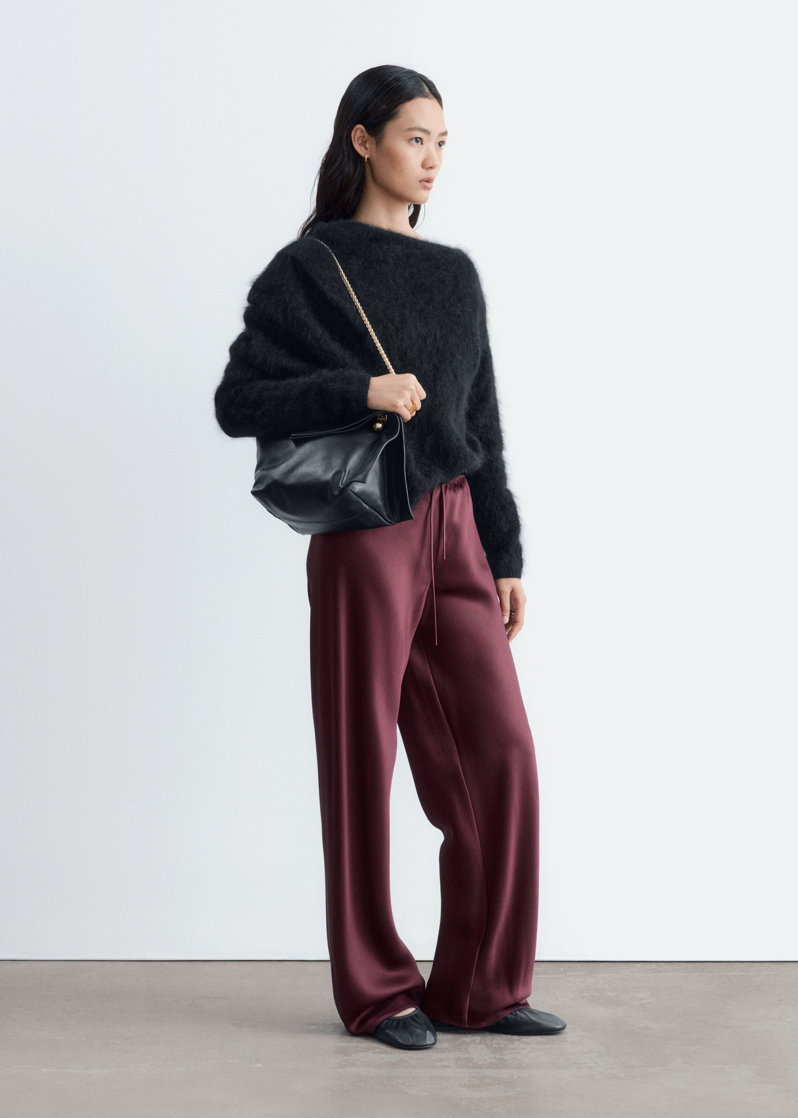 These burgundy satin trousers, featuring a relaxed wide-leg fit and an elastic drawstring waist, are worn by the model. The trousers are styled with a fuzzy black long-sleeve sweater, black flats, and a black leather shoulder bag.