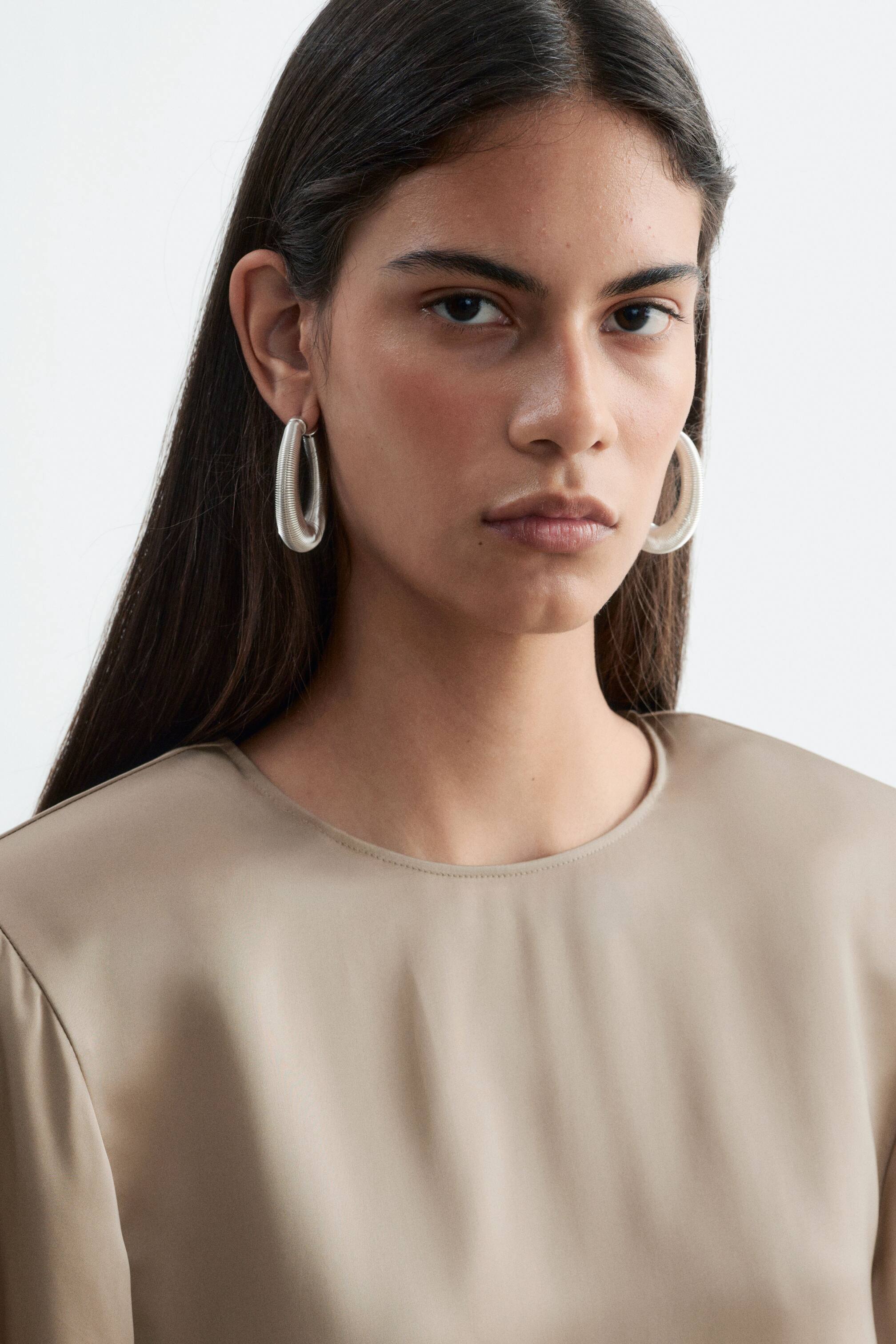 View larger image: A beige crew neck blouse featuring structured shoulders with subtle gathering is worn by the model. The blouse is styled with large, silver-toned hoop earrings.