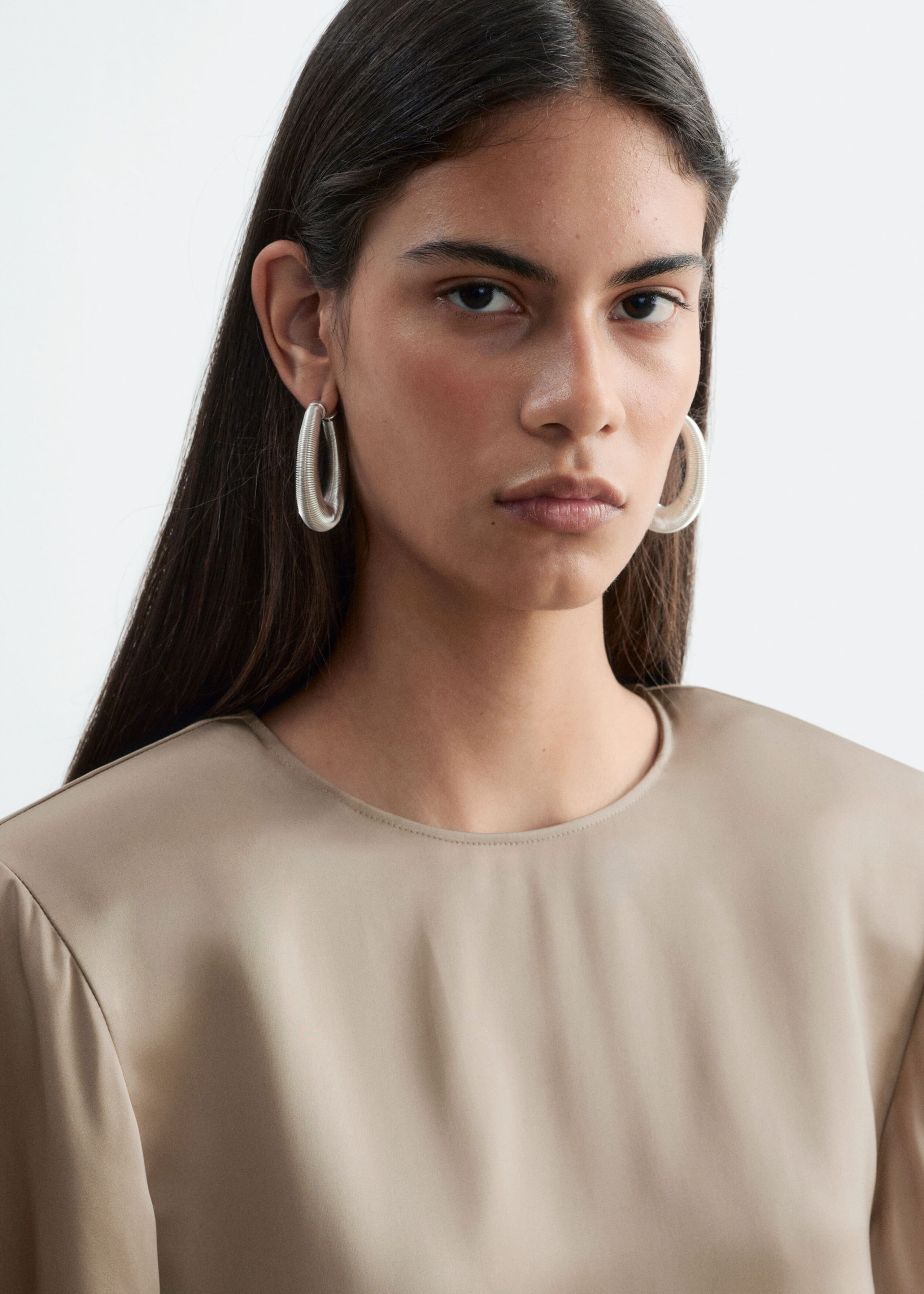 A beige crew-neck blouse featuring structured shoulders with subtle gathering is being worn by the model. The blouse is styled with large, silver-tone hoop earrings.