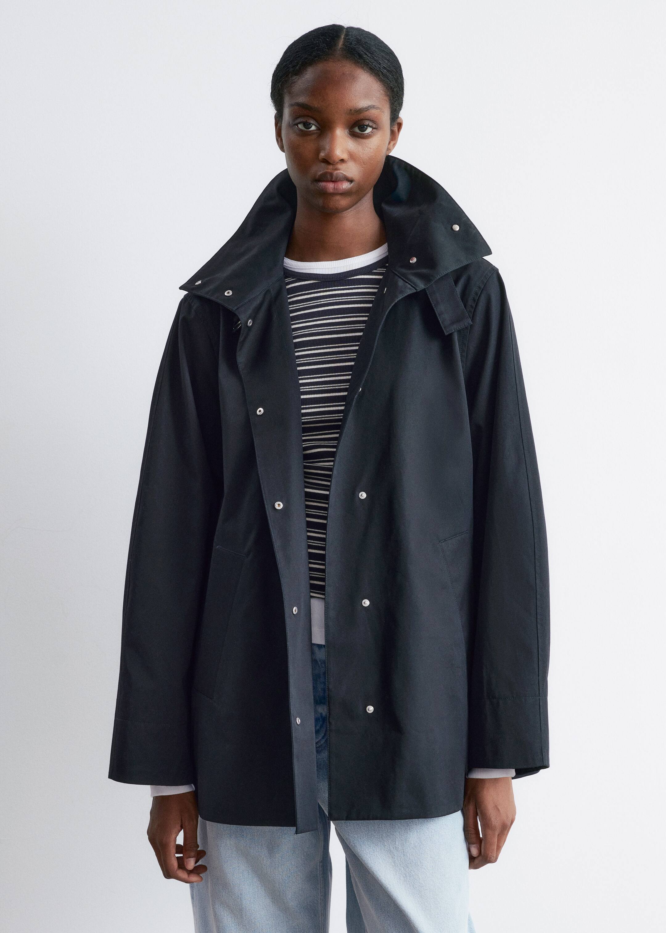 Grösseres Bild ansehen: Eine schwarze, hüftlange Jacke wird frontal präsentiert, offen getragen über einem horizontal gestreiften Oberteil und hellblauen Jeans. Die Jacke verfügt über einen hohen Stehkragen mit sichtbaren Druckknöpfen, lange gerade Ärmel und zwei sichtbare seitliche Schlitztaschen.