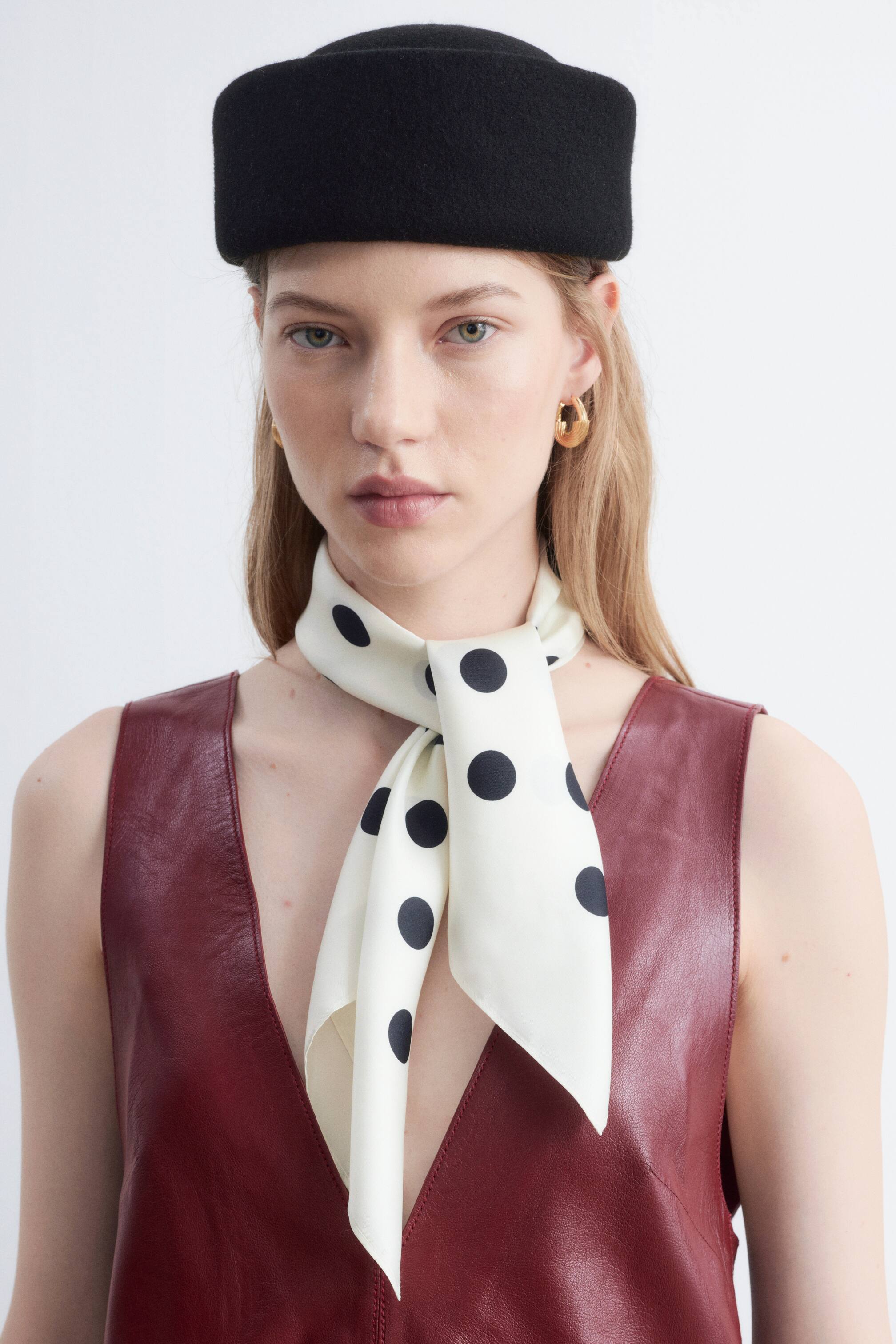 Grösseres Bild ansehen: Ein schwarzer Filzhut mit flacher Krone und kurzer, gerollter Krempe wird vom Model getragen. Ein cremefarbener Seidenschal mit schwarzen Polka Dots wird um den Hals gebunden getragen und über einem rötlich-braunen ärmellosen Kleidungsstück gestylt.