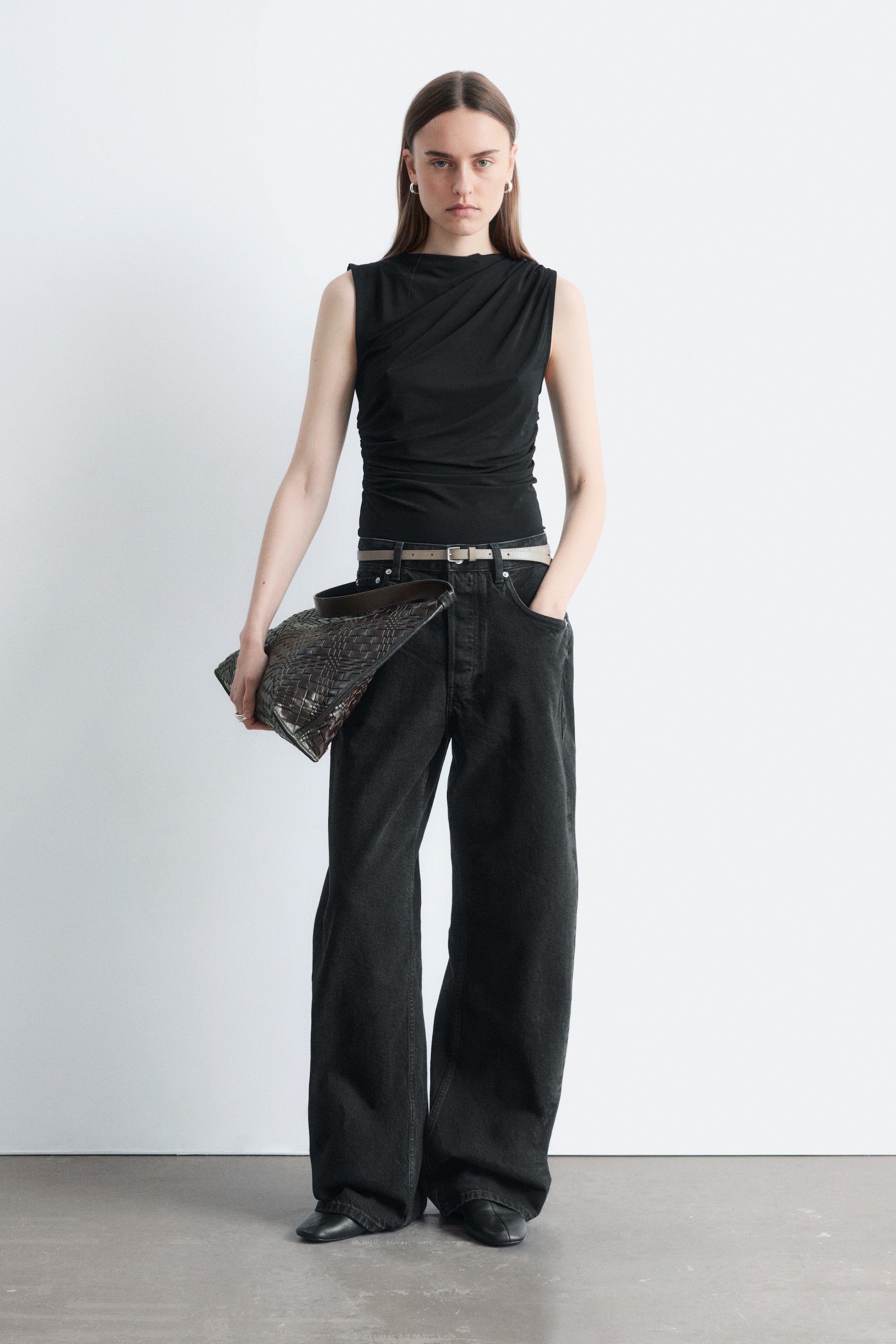 Ver imagen más grande: La modelo lleva puesto un top negro sin mangas con un escote barco y costuras fruncidas que crean un efecto drapeado. Está combinado con pantalones oscuros metidos por dentro.