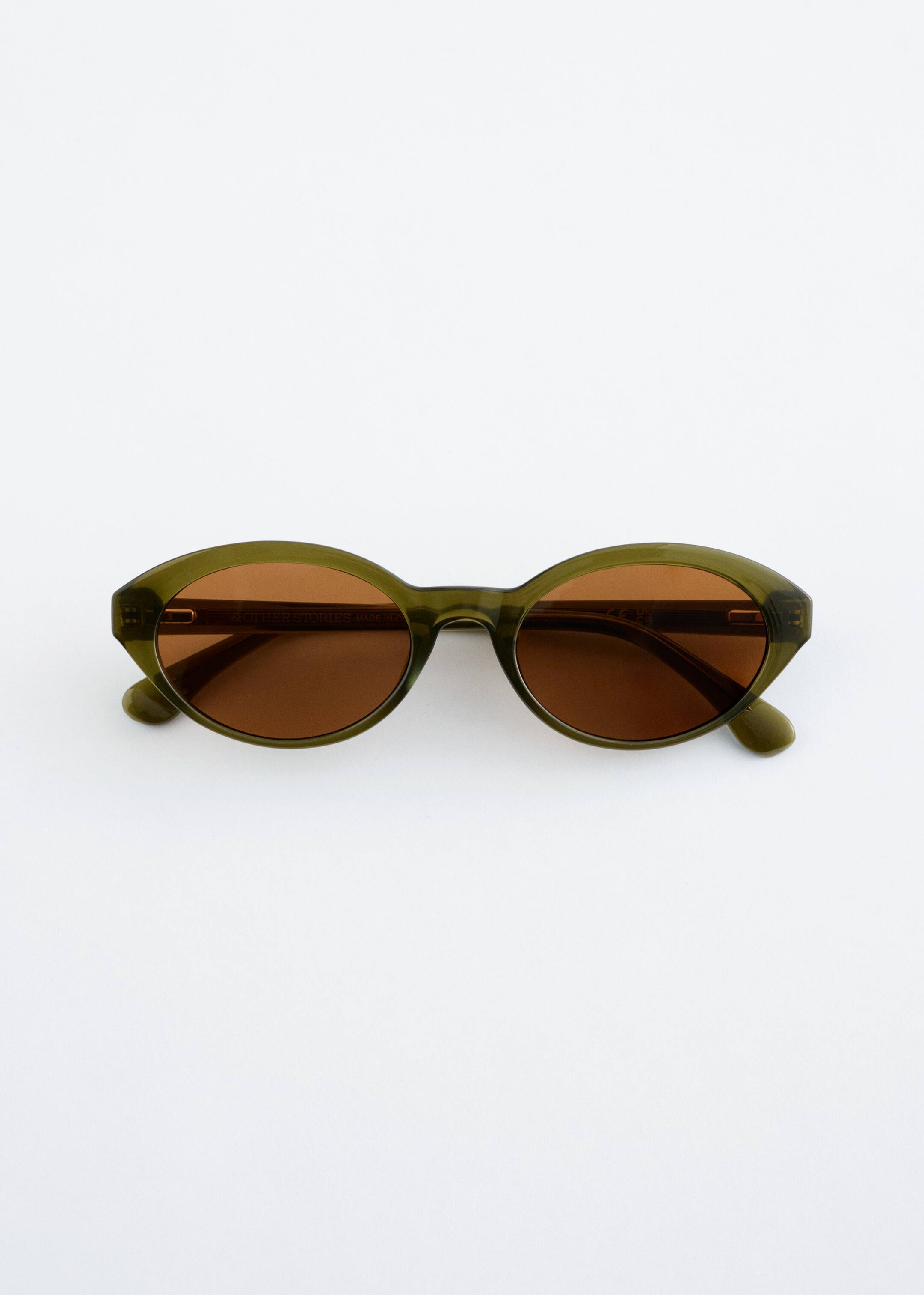 Zobrazit větší obrázek: Produkt je pár slunečních brýlí, směřující ČELEM. Mají průsvitné olivově zelené, oválné obroučky, které se jemně zužují po stranách a drží teplé hnědé čočky.