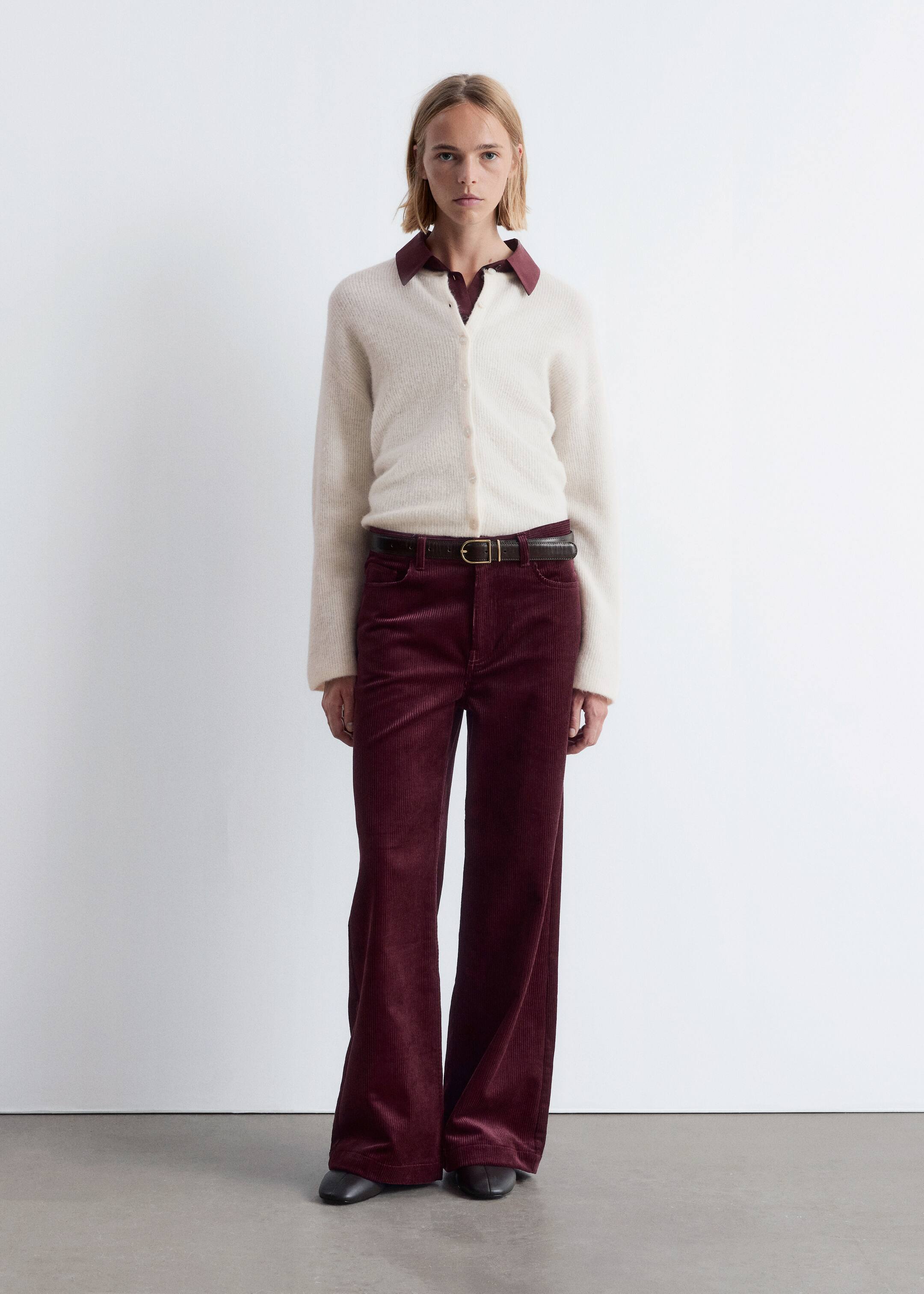 Visualizza immagine più grande: Il prodotto è una vista frontale a figura intera di un outfit che presenta un cardigan color crema, peloso, con scollo a V e chiusura a bottoni, indossato sopra una camicia con colletto rosso scuro. I pantaloni a coste larghe rosso scuro, con una silhouette a gamba larga, sono abbinati a una sottile cintura marrone scuro con fibbia in ottone, e scarpe chiuse scure completano il look.