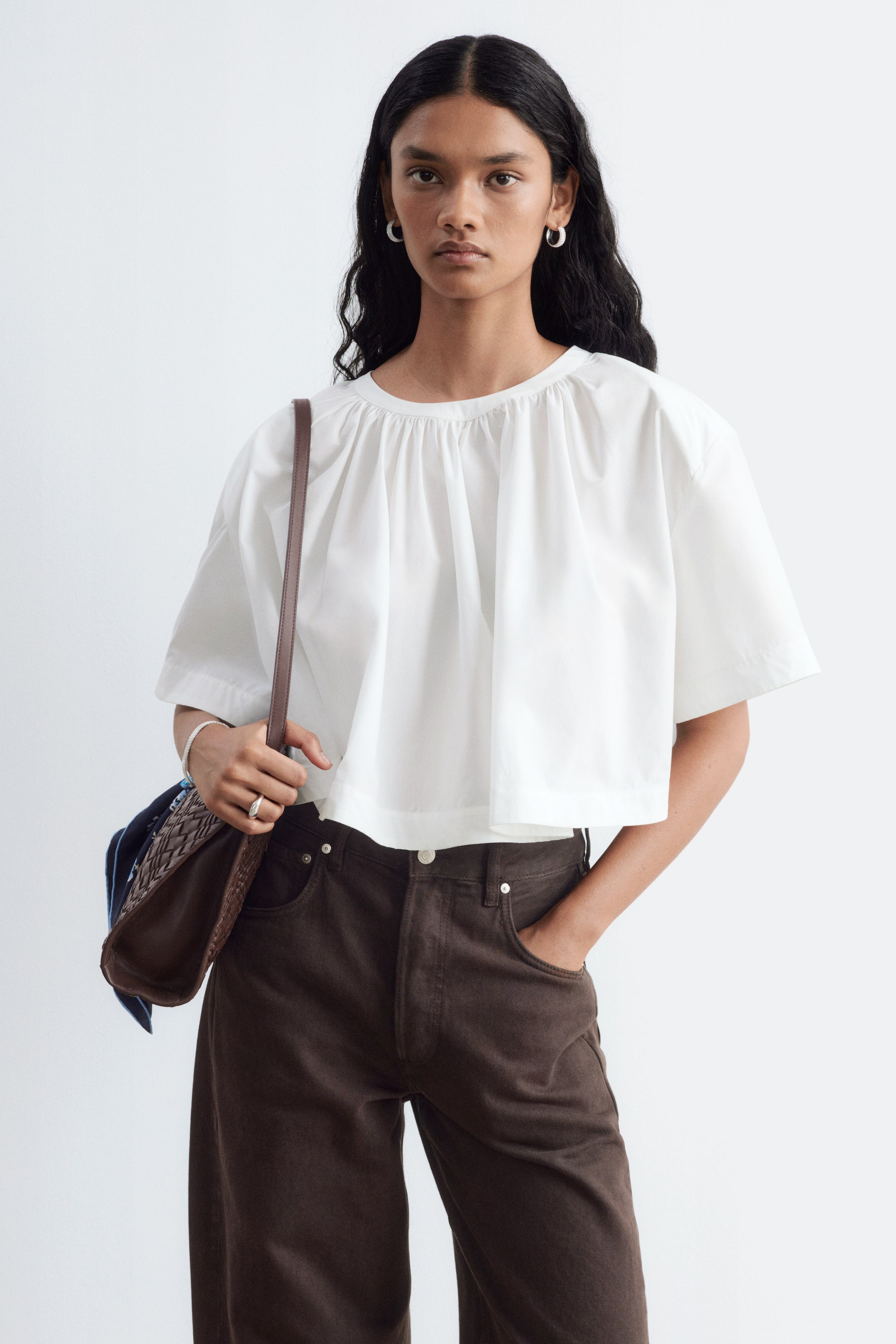 View larger image: A white gathered cotton crop blouse with a round, gathered neckline and wide, short sleeves is being worn by the model. The loose-fitting, cropped blouse is styled with dark brown trousers, while a brown shoulder bag is being carried on one side.