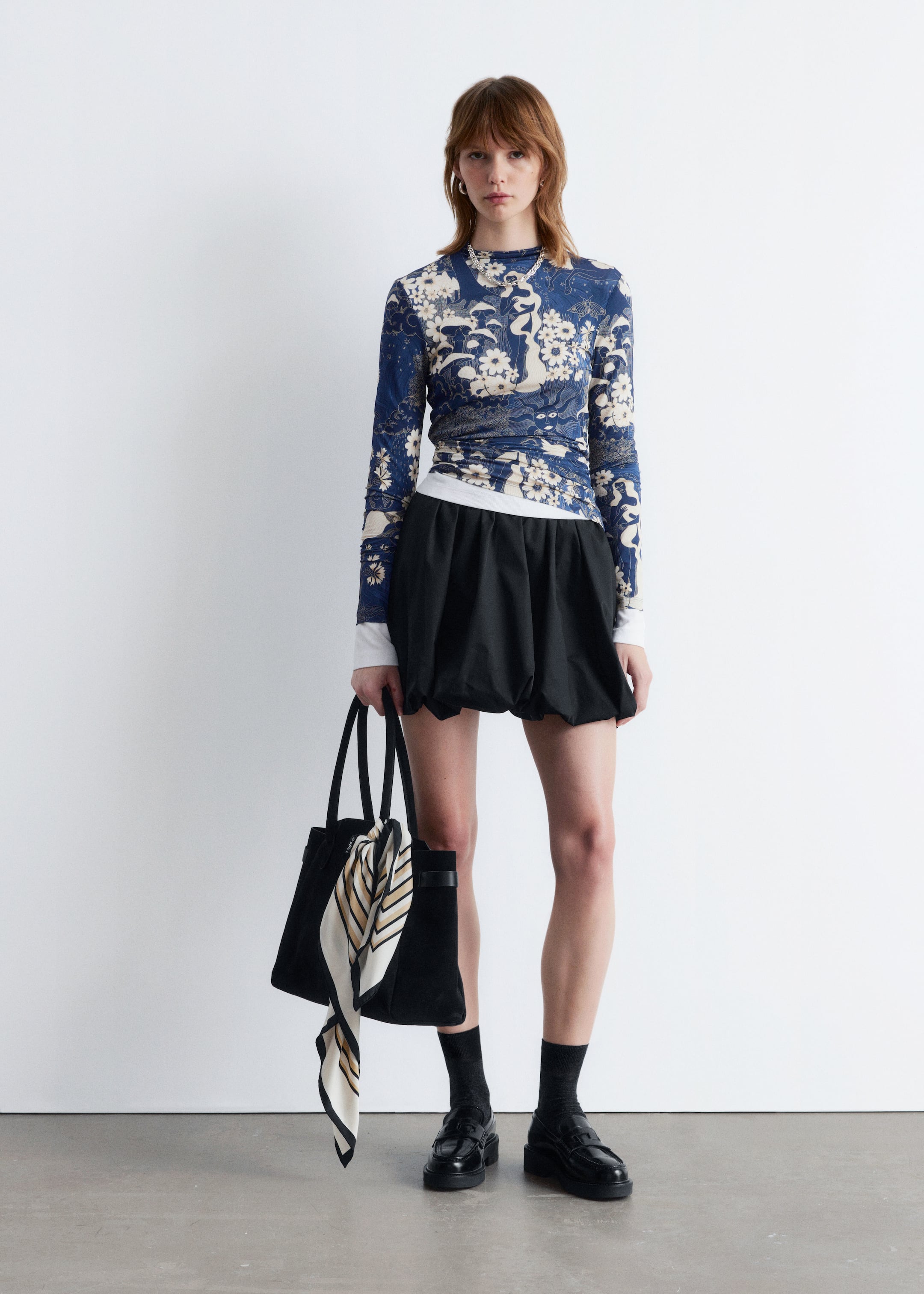 Größeres Bild anzeigen: Ein Model steht frontal und präsentiert ein langärmliges, hochgeschlossenes Oberteil in einem blauen und weißen Blumen- und abstrakten Druck, das über einem weißen langärmligen Oberteil getragen wird, kombiniert mit einem schwarzen voluminösen kurzen Rock mit geraffter Taille und einem gewellten Blasen-Saum. Sie trägt eine schwarze Tragetasche mit einem gestreiften Schal, der am Griff befestigt ist, und schwarze klobige Loafer mit schwarzen Socken.