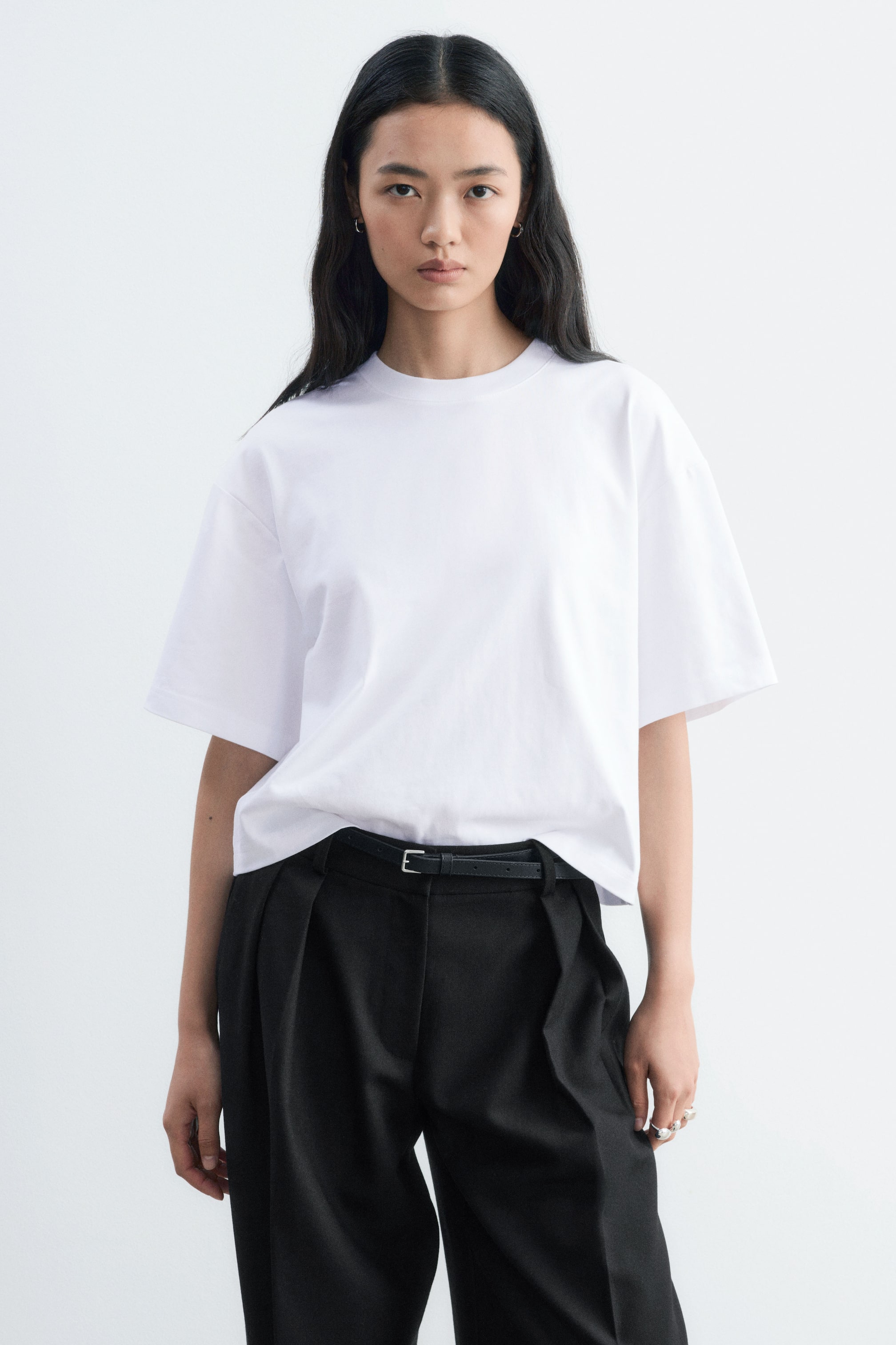 Visualizza immagine più grande: Una t-shirt bianca dalla vestibilità rilassata, con classico girocollo e maniche corte ampie, indossata dalla modella. La t-shirt è infilata morbida sul davanti in pantaloni neri stretti in vita da una sottile cintura nera.