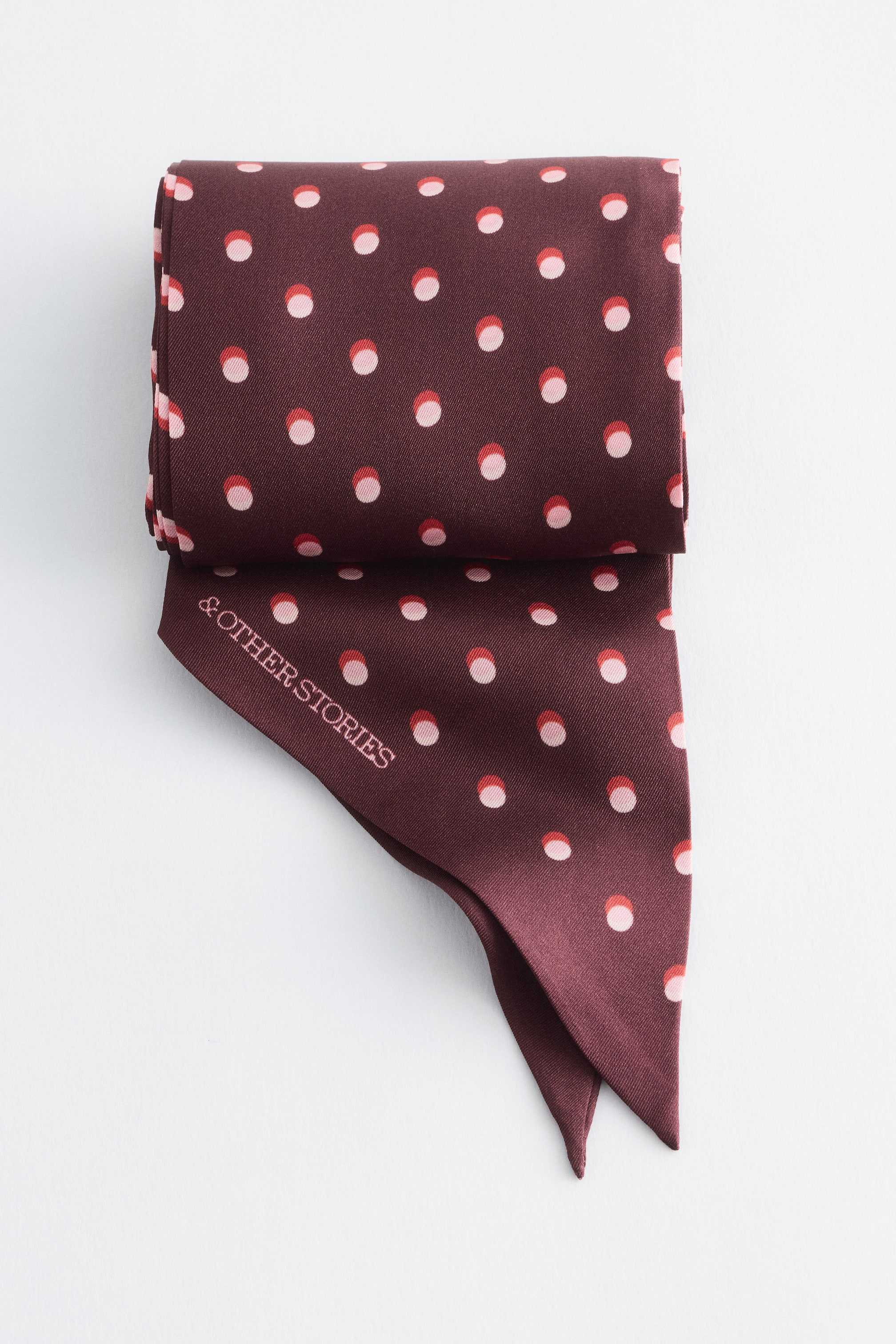 Grotere afbeelding bekijken: Een smalle bordeauxrode zijden sjaal met een all-over patroon van tweekleurige roze en rode polkadots wordt bovenaan opgevouwen getoond, waarbij de puntige uiteinden naar beneden hangen. De merknaam "& OTHER STORIES" staat in lichtroze gedrukt langs een van de diagonale randen.