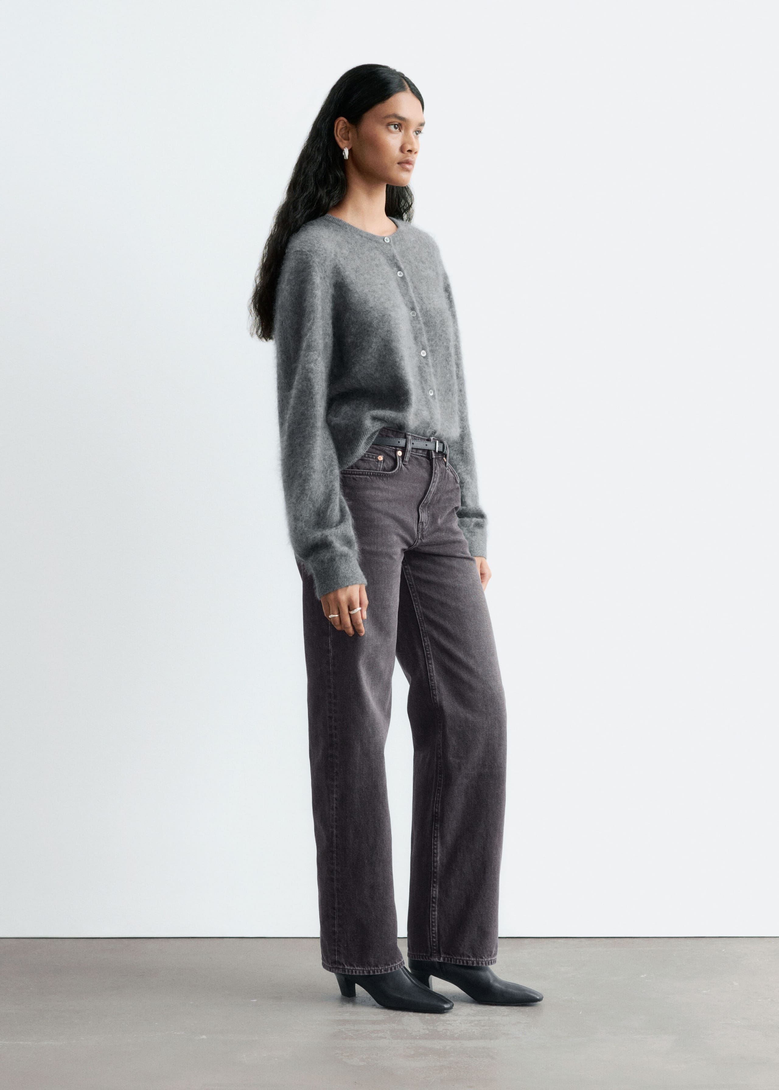 Straight-leg, mid-rise jeans in washed dark gray denim are worn by the model, who is standing in a three-quarter profile. The jeans are styled with a thin black belt, a partially tucked-in gray fuzzy cardigan, and black pointed-toe heeled boots.