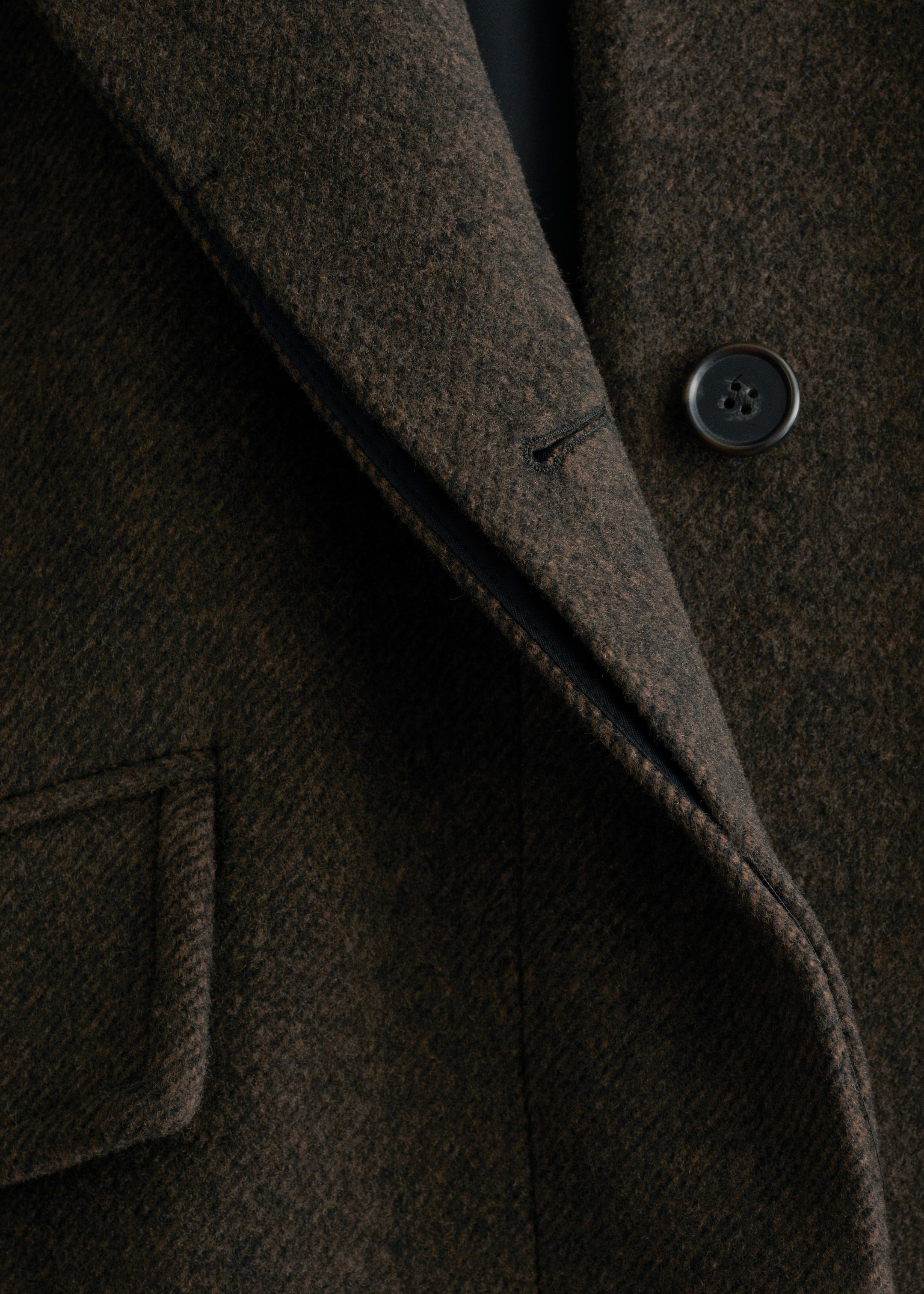 View larger image: TOP: A dark brown wool blend coat is being shown in a close-up, featuring a single visible dark button and an adjacent buttonhole on its front. A slanted flap pocket is also visible below.