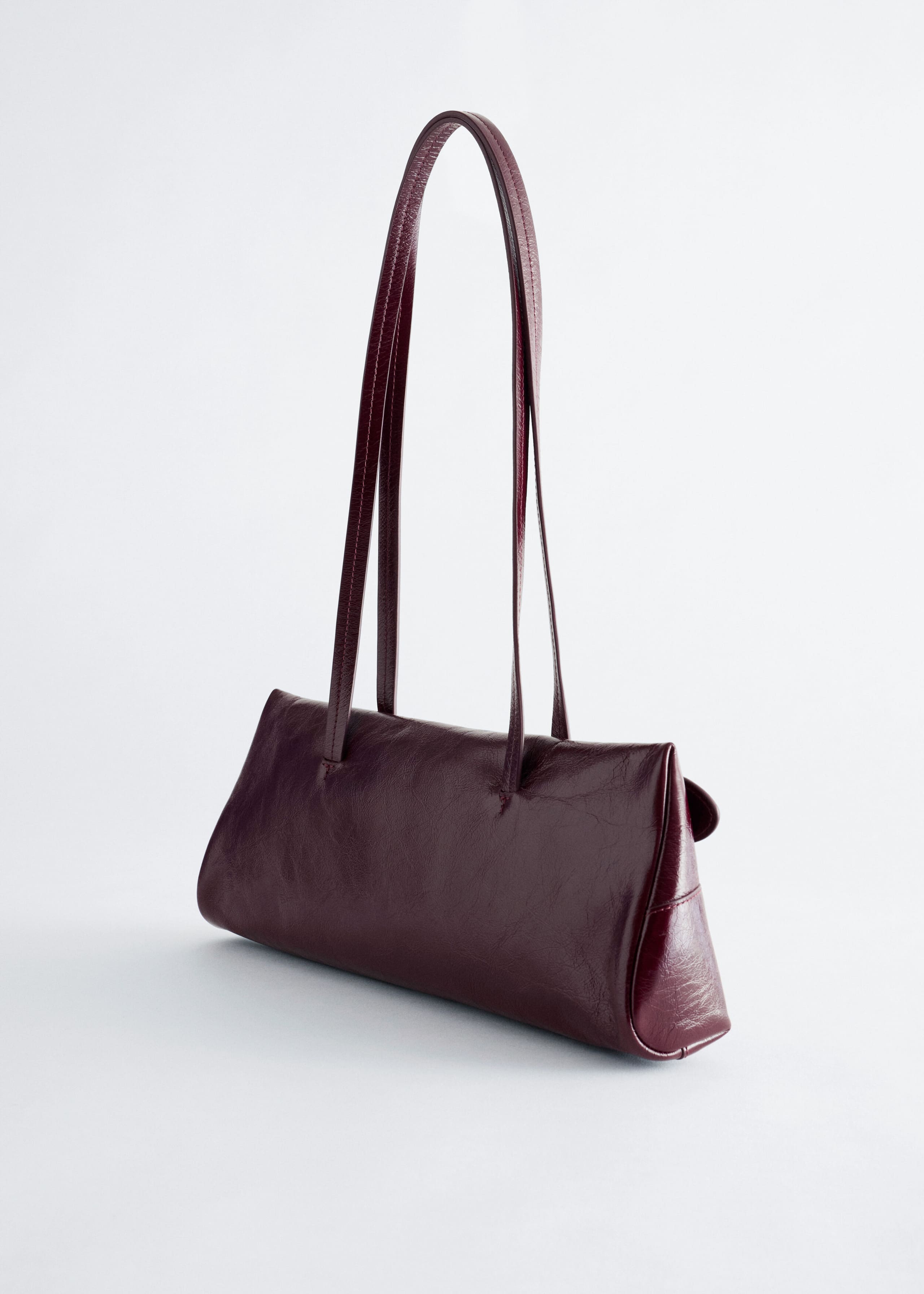 A burgundy leather shoulder bag with a low-profile rectangular silhouette is posed at an angle, showcasing its elongated, slim shoulder straps. The bag features a soft, slightly textured leather finish with neat stitched detailing.