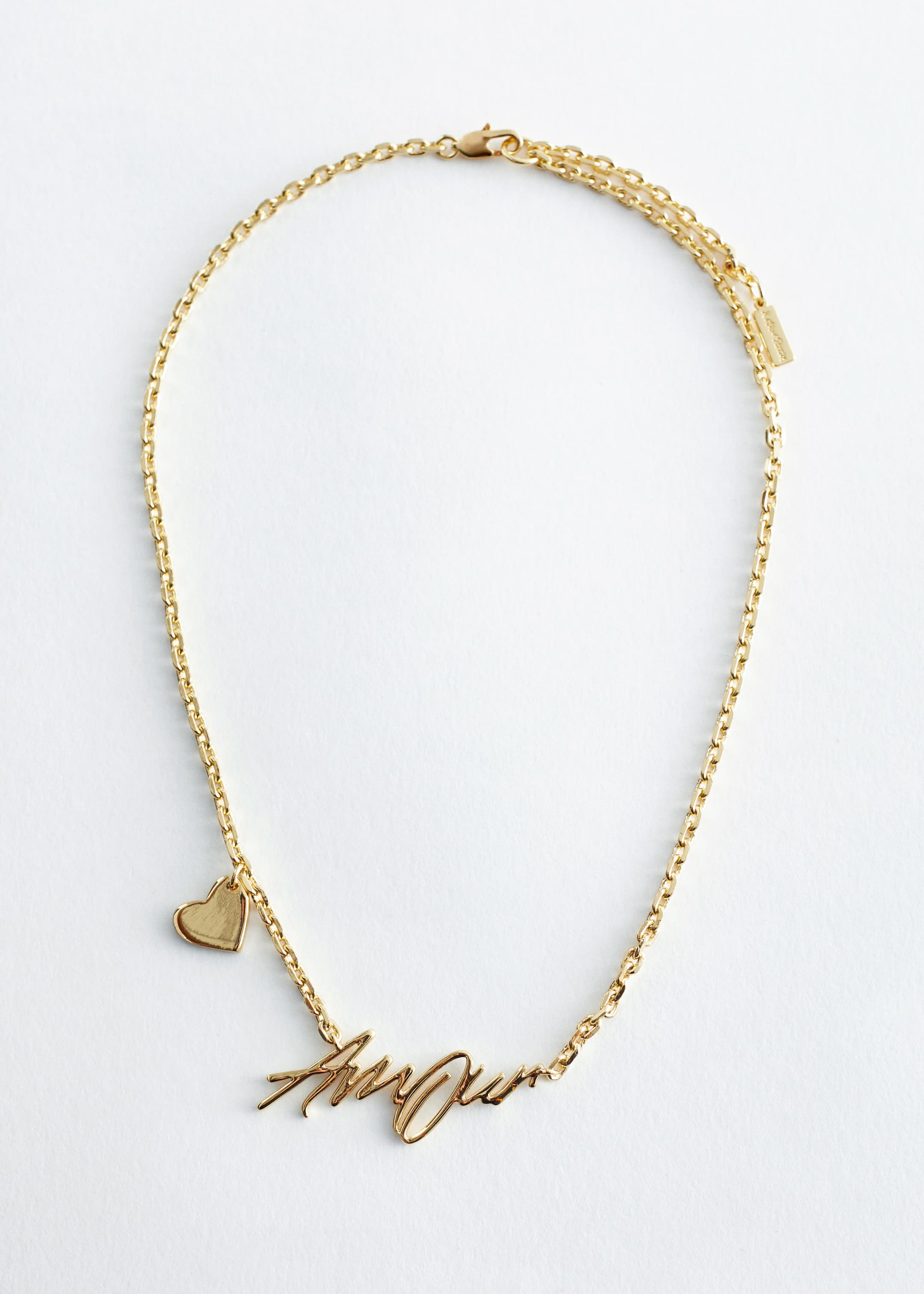 View larger image: A polished brass necklace is featuring a delicate rolo chain, with a text-motif pendant spelling "Amour" positioned at the FRONT. A heart charm is accenting one side of the chain, and the necklace is finished with an adjustable length and a trigger clasp.