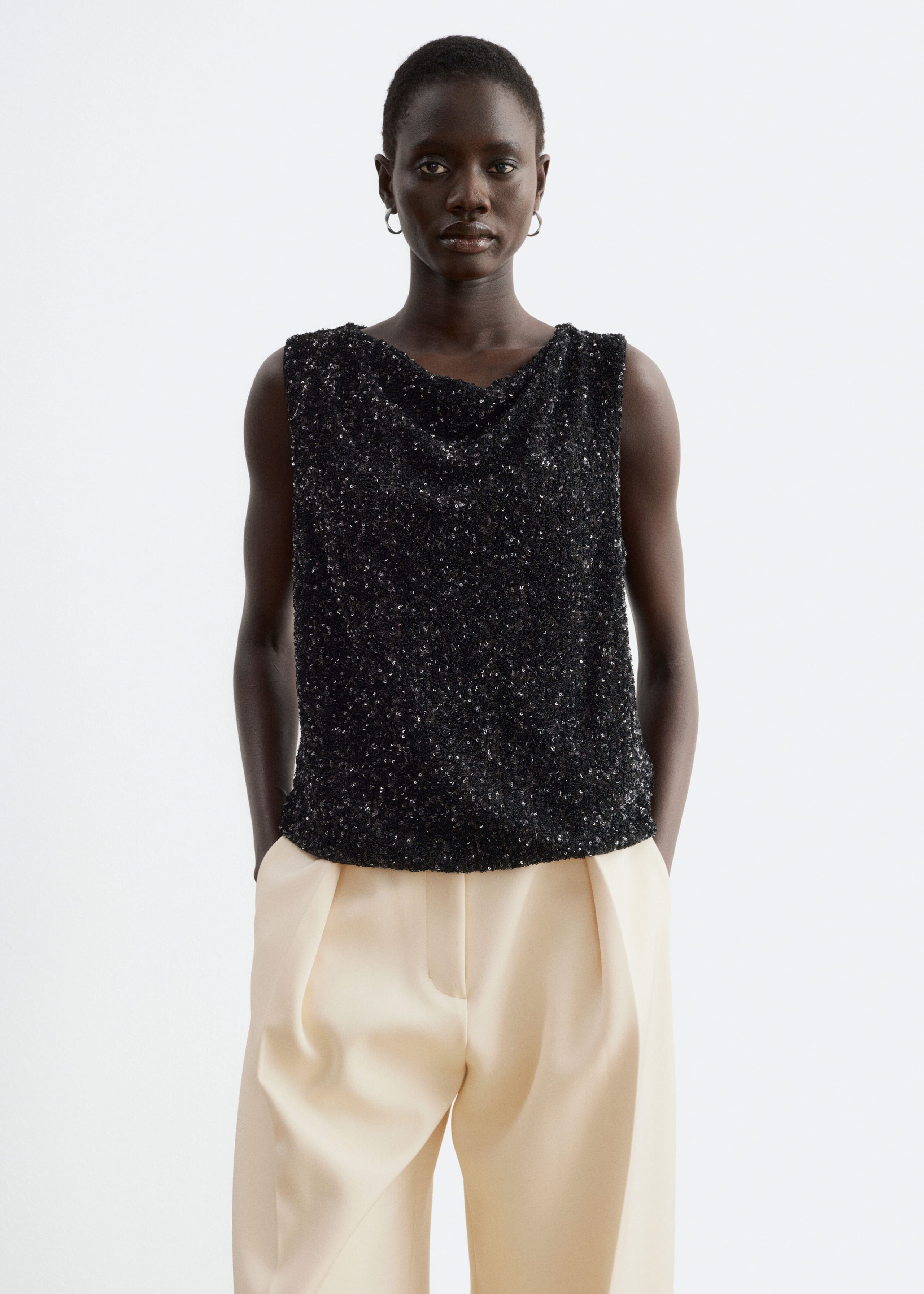 A black, sleeveless sequinned top featuring a draped neckline is being worn by the model. The top is styled with cream-coloured, pleated trousers.