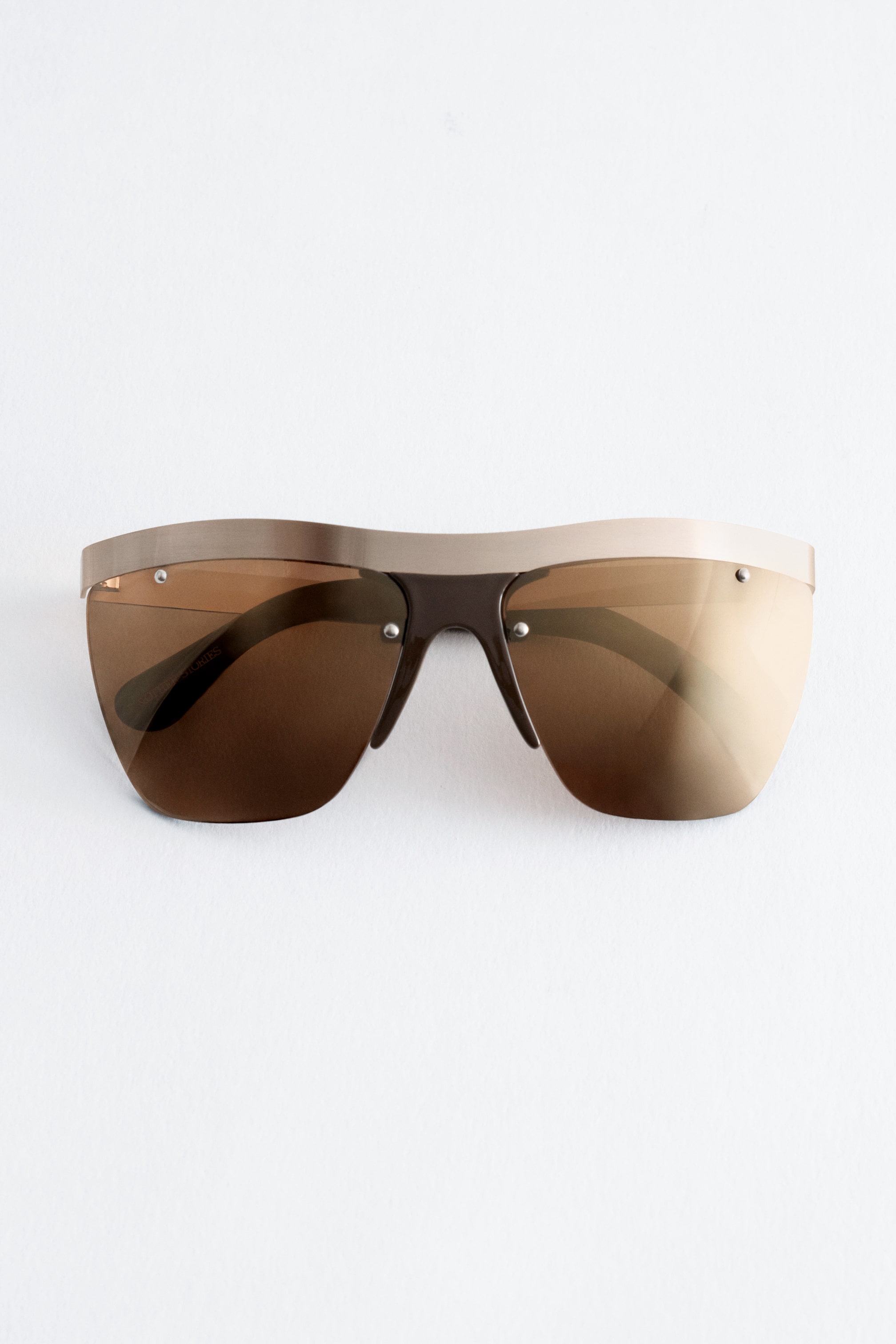 View larger image: These shield sunglasses, viewed from the front, feature semi-translucent brown lenses topped with a wide, metallic gold-toned brow bar. Small metallic studs secure the lenses to the frame, with dark brown temples visible through the tinted lenses.