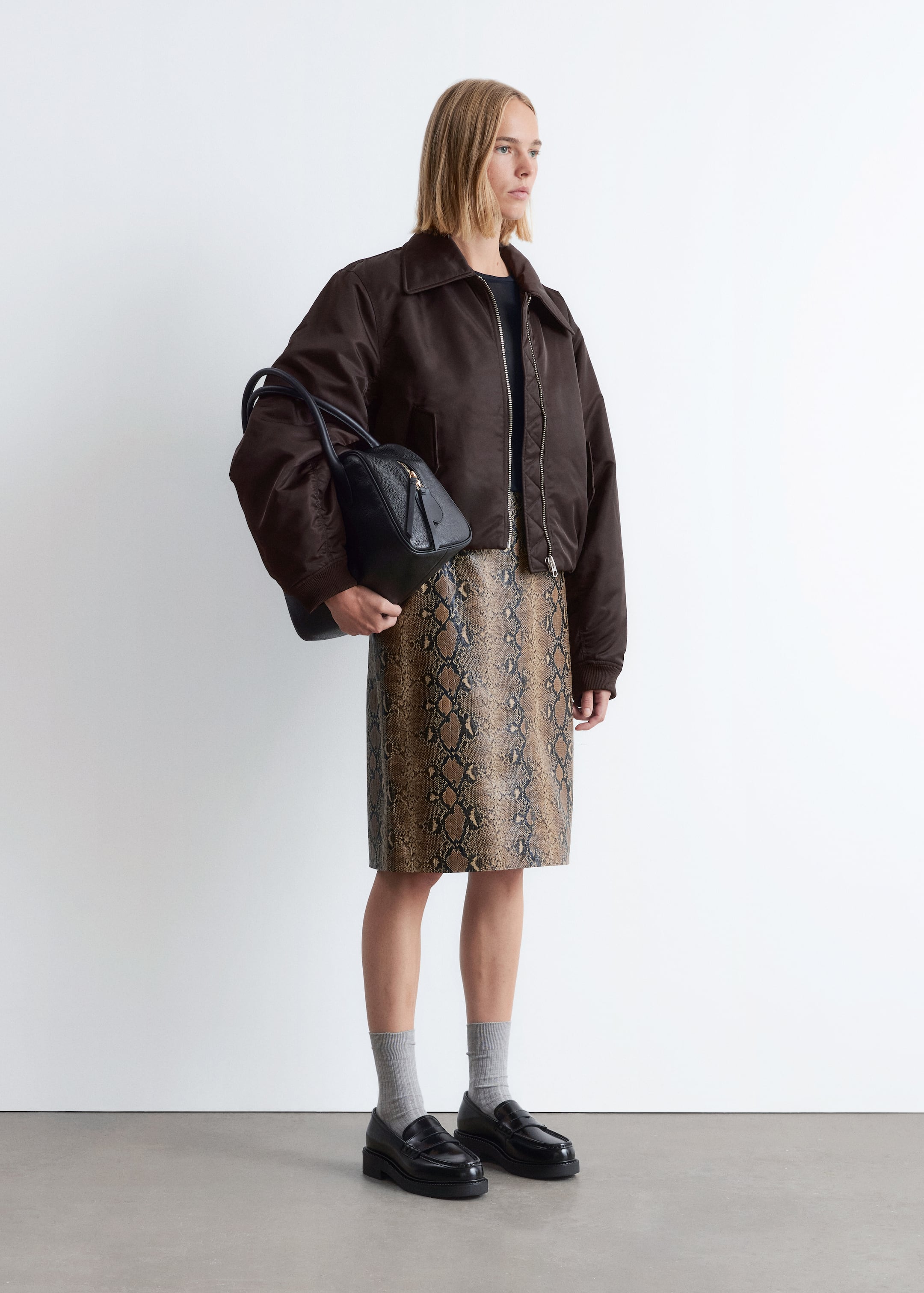 Ingrandisci l'immagine: Un ensemble è presentato su una persona, visto da un leggero angolo laterale. Presenta un bomber oversize marrone con un ampio colletto e polsini a coste, indossato sopra una t-shirt nera. Una gonna dritta di lunghezza midi con una stampa pitonata marrone e nera si estende fino alle ginocchia. Una borsa strutturata nera con due manici superiori e una cerniera dorata è tenuta in mano, e mocassini neri chunky completano il look, abbinati a calzini a coste grigio chiaro.