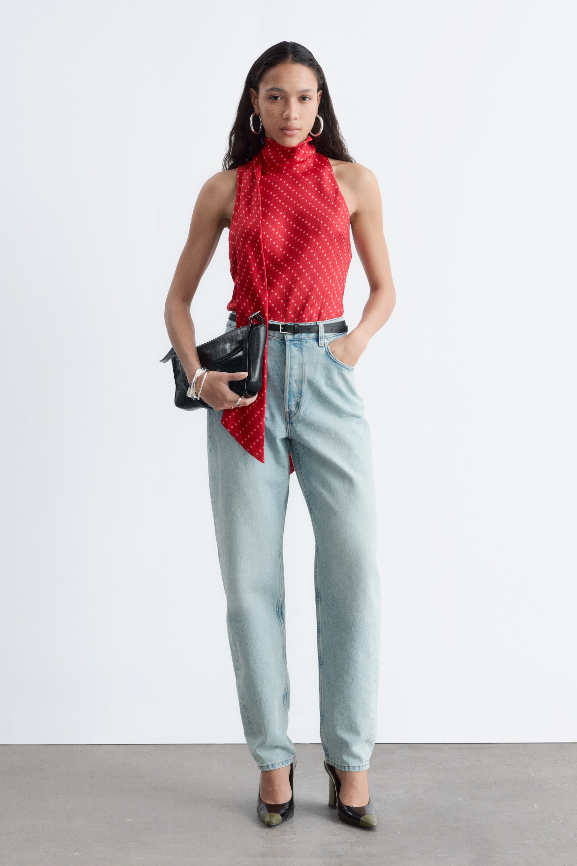 View larger image: A red sleeveless top, featuring a high tie-neck and a white polka dot pattern, is tucked into light wash blue straight-leg jeans with a black belt. A black structured clutch bag is being held, and black high-heeled pumps with green pointed toes are being worn.