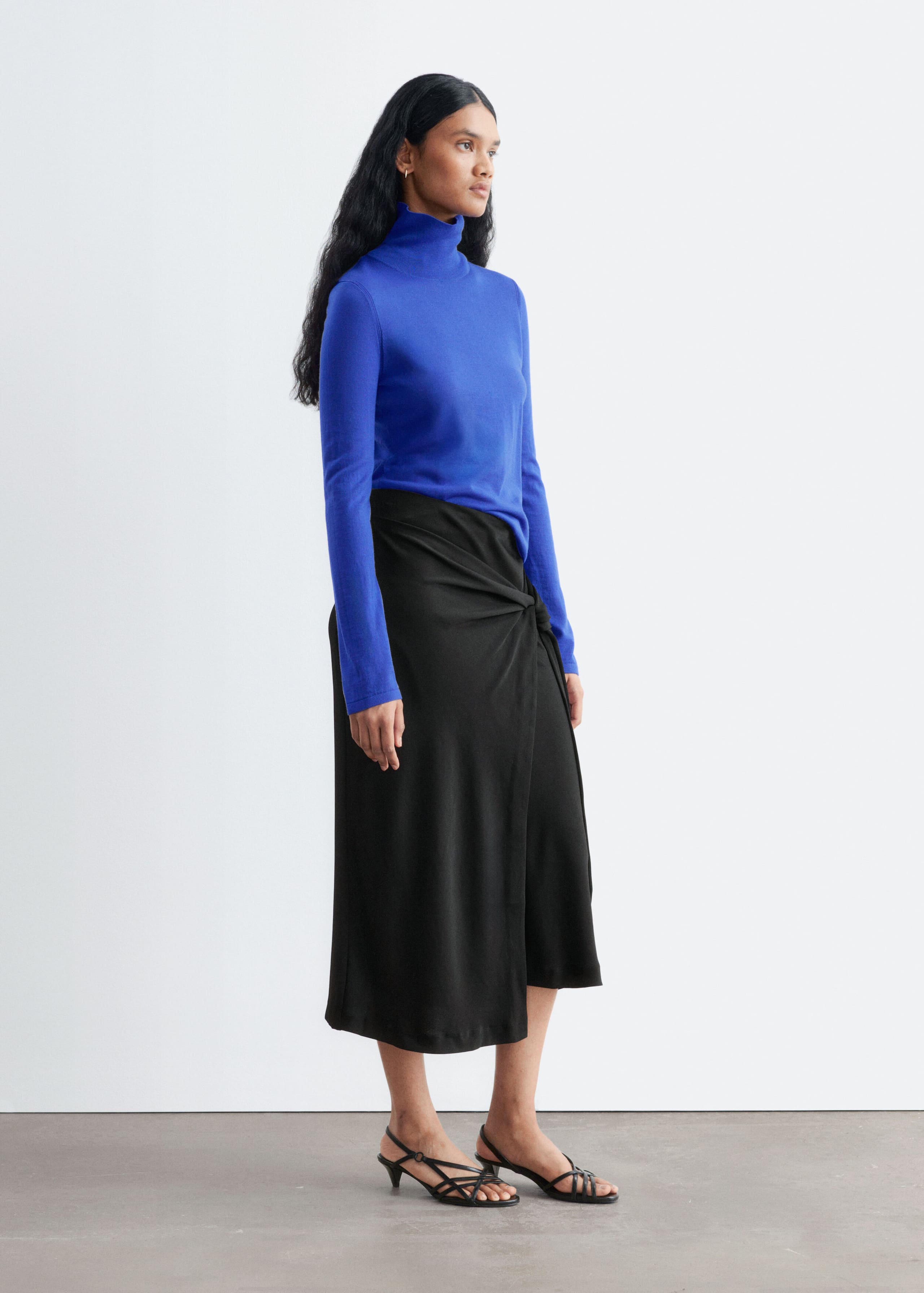 Een blauw Merino-wollen coltruitje met hoge kraag en lange mouwen wordt gedragen door het model. Het topje is gestyled en ingestopt in een zwarte gedrapeerde midi-rok met een knoopdetail in de taille.