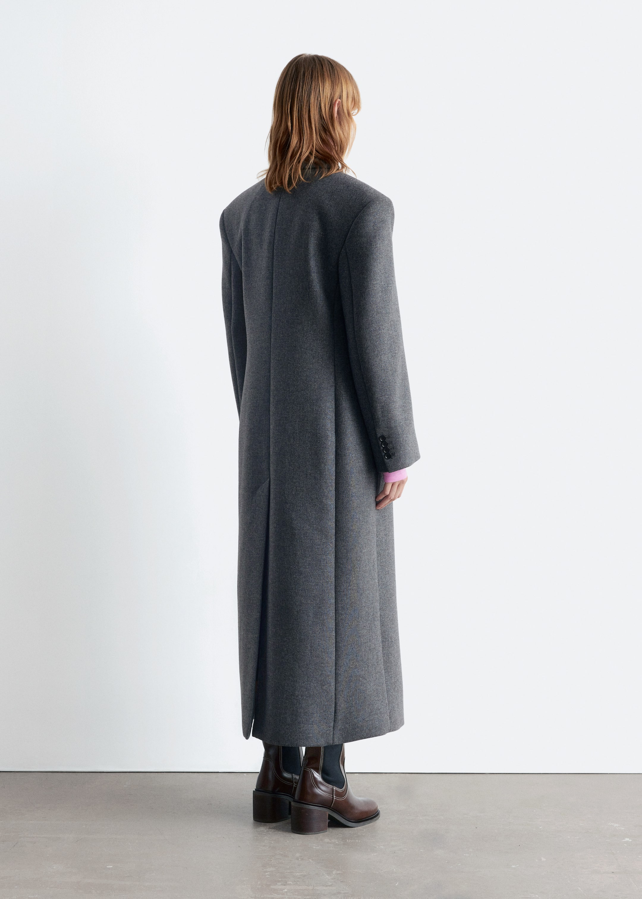 Agrandir l'image: Un long manteau gris foncé est montré de dos, avec des épaules structurées, une couture verticale centrale et une seule fente au bas du dos. Ses longues manches affichent trois boutons visibles sur les poignets, tandis qu'un haut rose à manches longues est visible en dessous et des bottes brunes apparaissent sous l'ourlet.