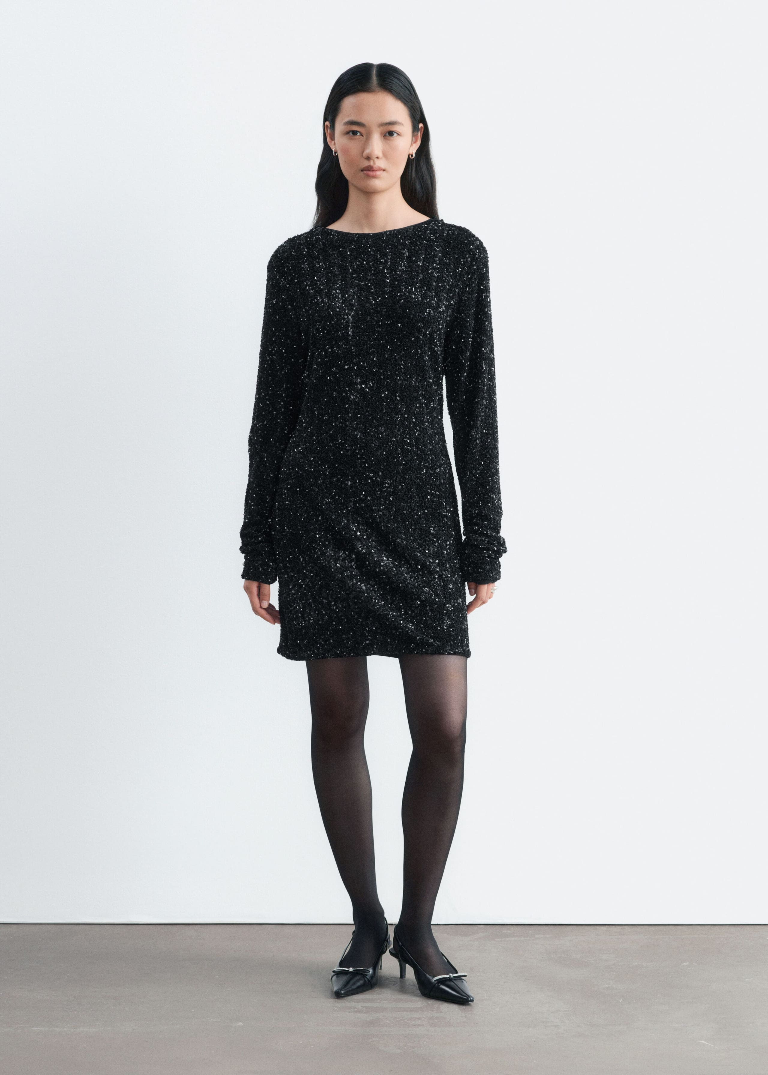 A black sequined mini dress with long sleeves and a high, rounded neckline is being worn by the model. The dress is styled with sheer black tights and black pointed-toe heels.