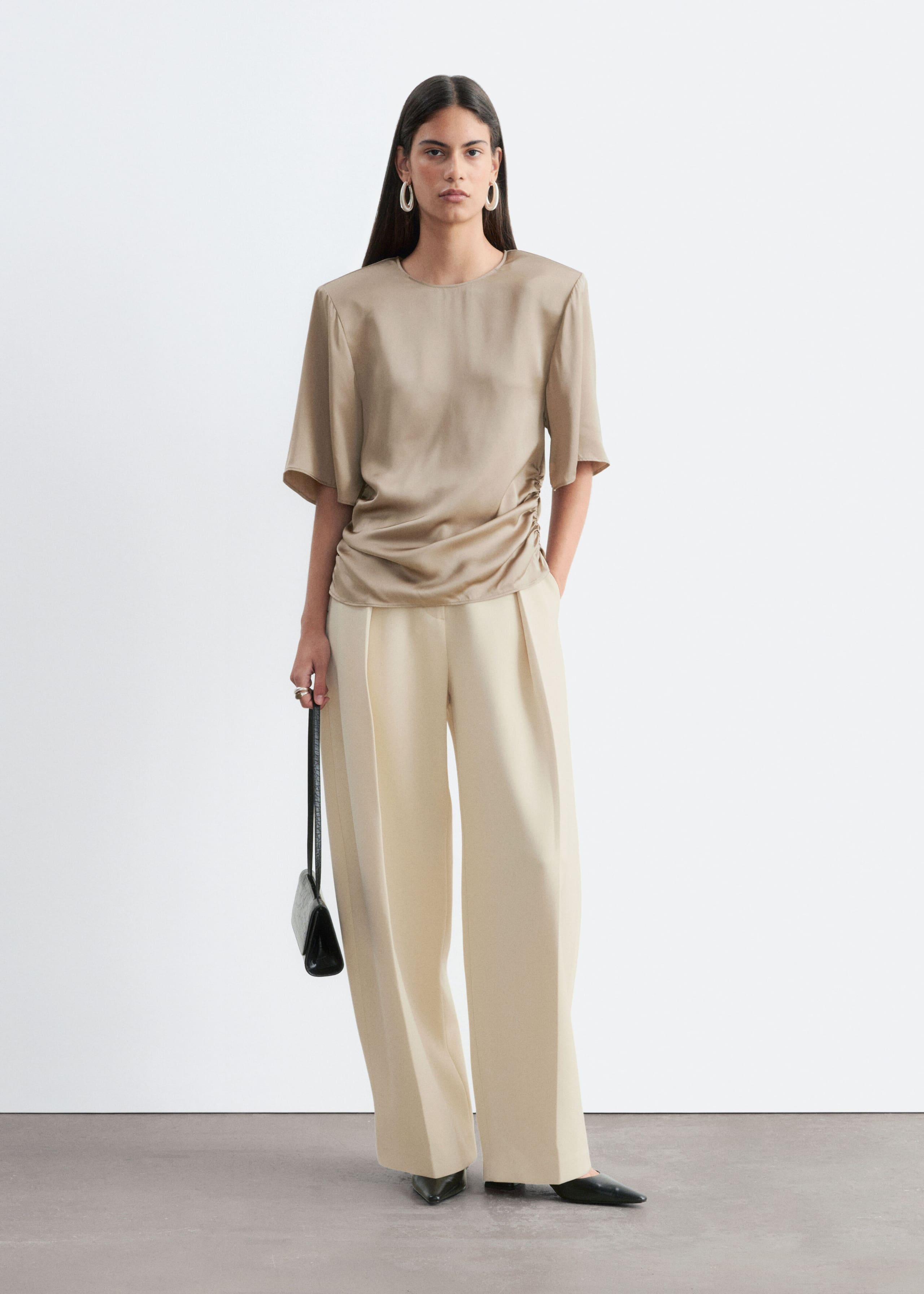 A beige, slim-fit blouse with soft shoulder pads, short sleeves, and gathered side pleats is worn by the model. The blouse is styled with cream-colored wide-leg trousers and a small black handbag carried in one hand.