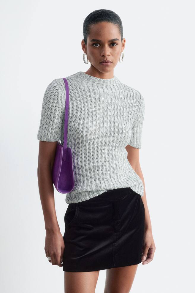La modelo lleva un top de punto metalizado con una ligera textura acanalada en color plateado, cuello redondo alto y mangas cortas. El top se combina con una falda de terciopelo negro y un bolso de hombro morado llevado al hombro. - 1
