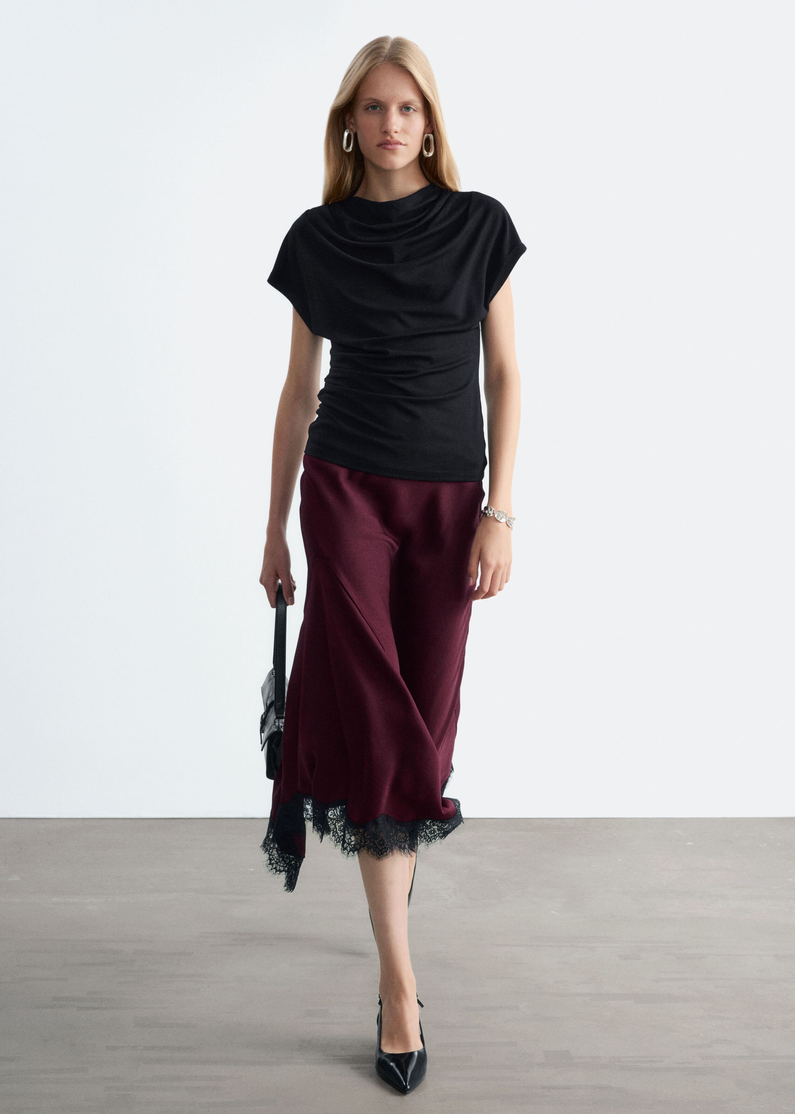 A burgundy flared midi skirt featuring an asymmetrical hemline detailed with black lace is being worn by the model. The skirt is styled with a draped black top and black heels.