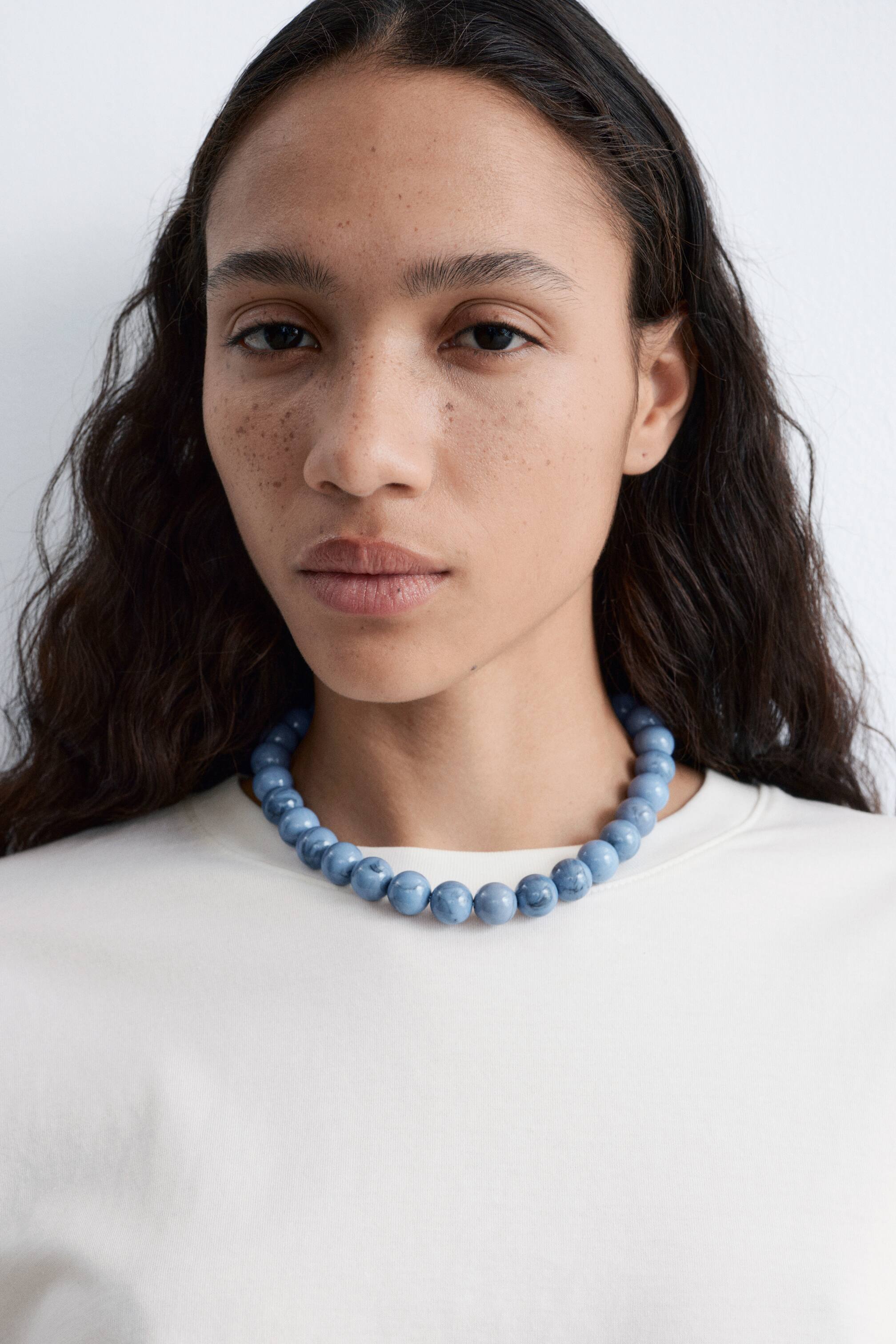 Se større billede: En kort halskæde med blå stenperler bæres af modellen og hviler på en hvid top.
