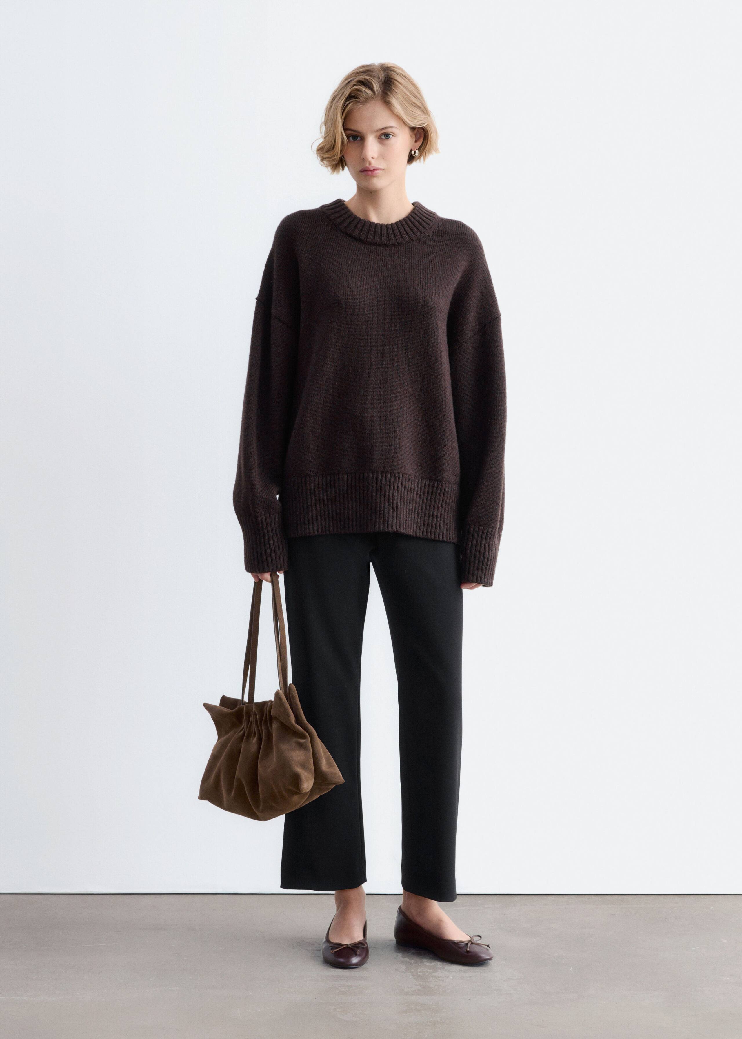 The model wears an oversized dark brown wool sweater featuring a ribbed crew neck, dropped shoulders, and wide ribbed cuffs and hem. The sweater is styled with black straight-leg trousers, dark brown flat shoes, and a brown suede handbag held in one hand.