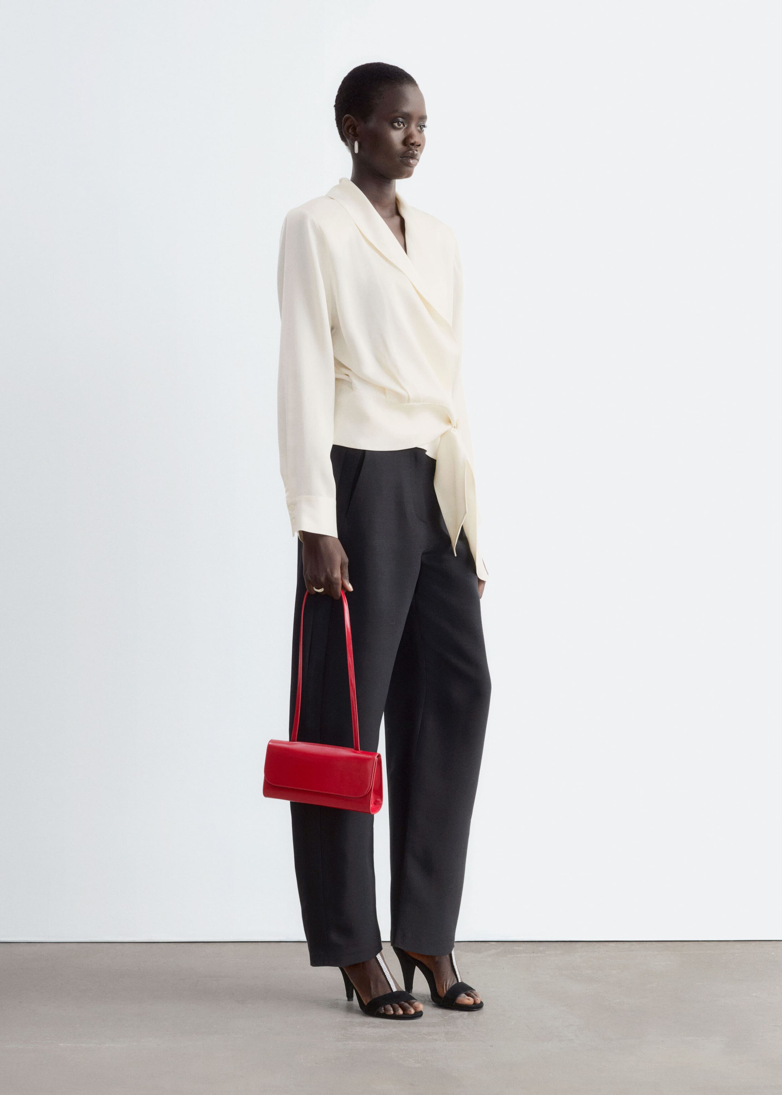 La modella indossa una blusa avvolgente color crema con morbido colletto a scialle, maniche lunghe e chiusura in vita con laccetto annodato sul davanti. La blusa è abbinata a pantaloni neri a gamba larga, mentre nella mano la modella porta una piccola borsa rossa a tracolla.