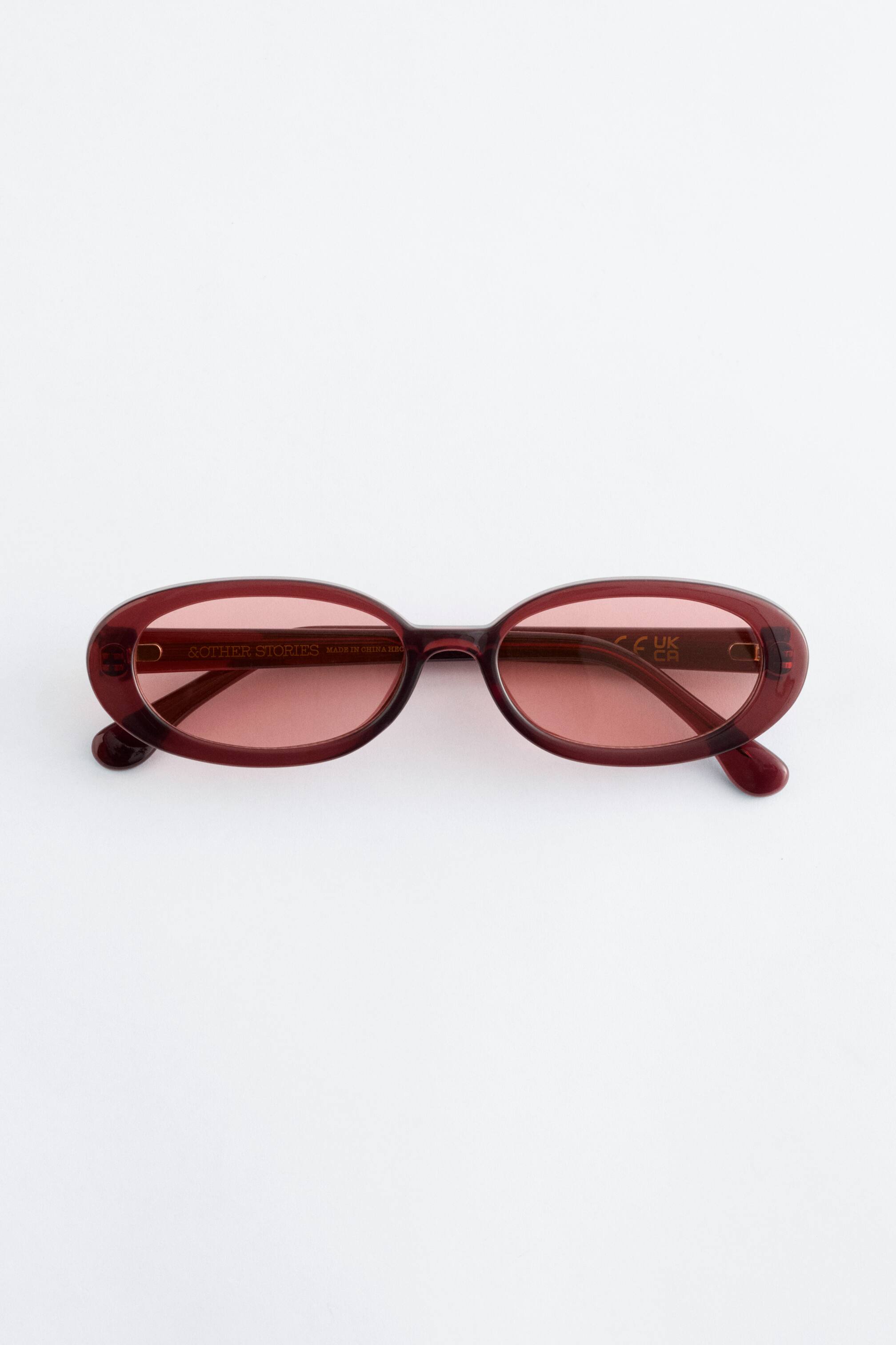 Vezi imaginea mai mare: O pereche de ochelari de soare burgundy este prezentată din față, având o ramă ovală mică și lucioasă, cu lentile nuanțate în roz tonal și brațe subțiri.