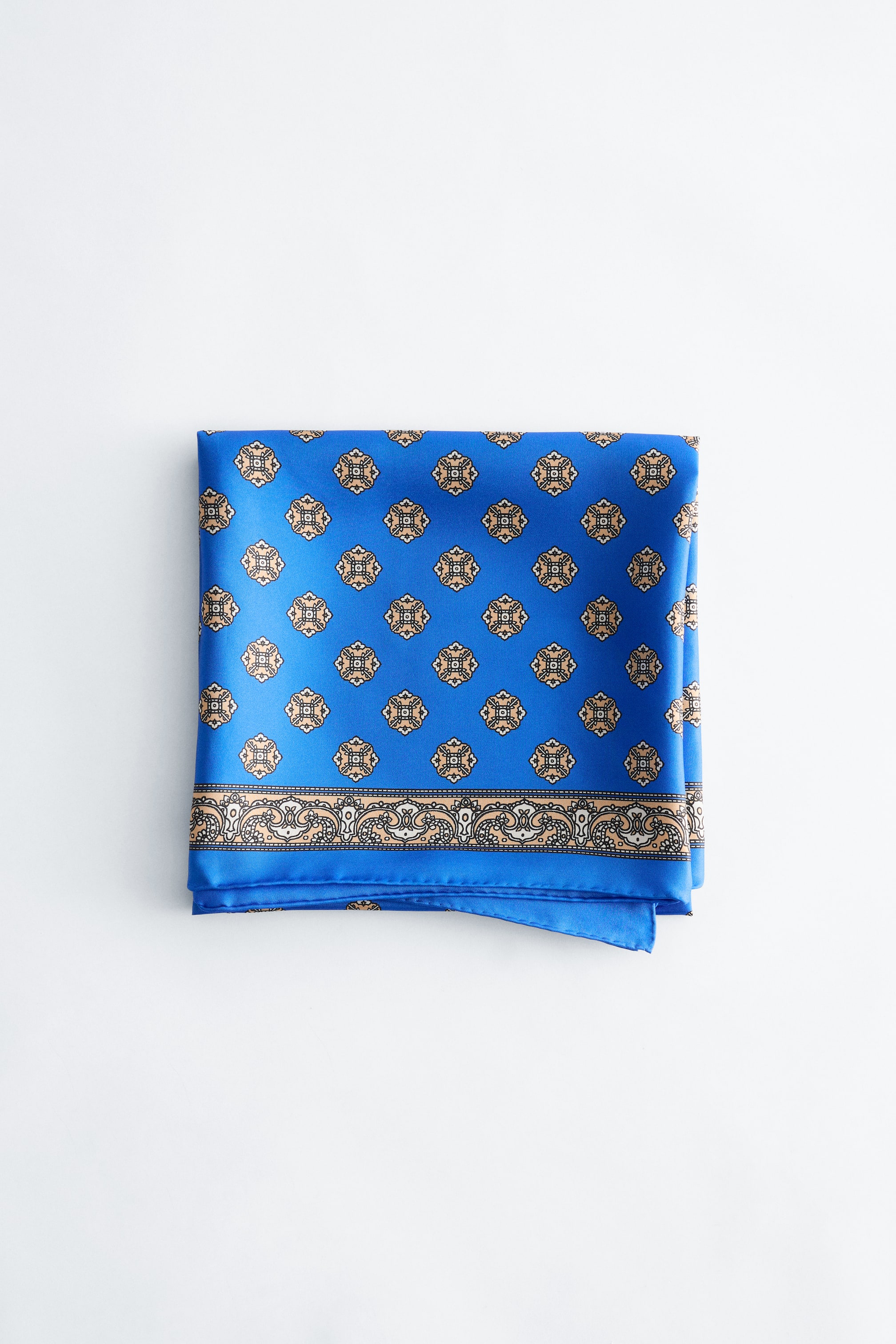 View larger image: A blue scarf is folded flat, facing front, displaying a repeating pattern of light brown, black and white square motifs across its surface and a wider decorative border with intricate scrollwork in matching colours along its bottom edge.