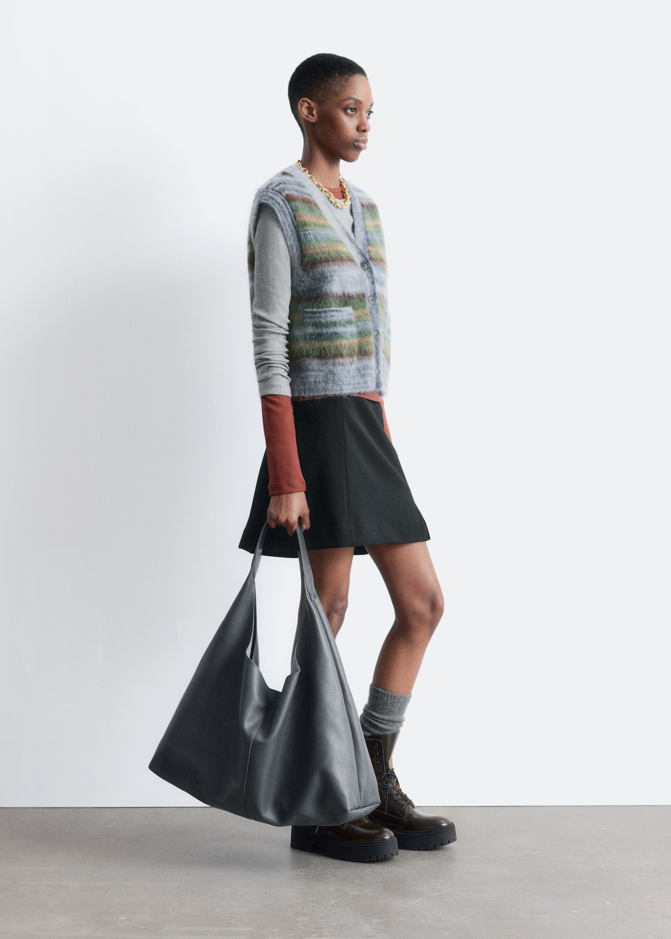 Visualizza immagine più grande: Un abbigliamento completo viene presentato da un angolo laterale, caratterizzato da un gilet a maglia pelosa con scollo a V e bottoni sul davanti con righe orizzontali in grigio, verde, arancione e marrone, stratificato su un top a maniche lunghe grigio chiaro con polsini marrone rossiccio e una collana a catena robusta dorata. Il look si completa con una mini gonna nera a trapezio, calzini grigi a coste, stivaletti stringati marrone scuro con suole robuste e una grande borsa a tracolla grigio scuro testurizzata e morbida portata a mano.