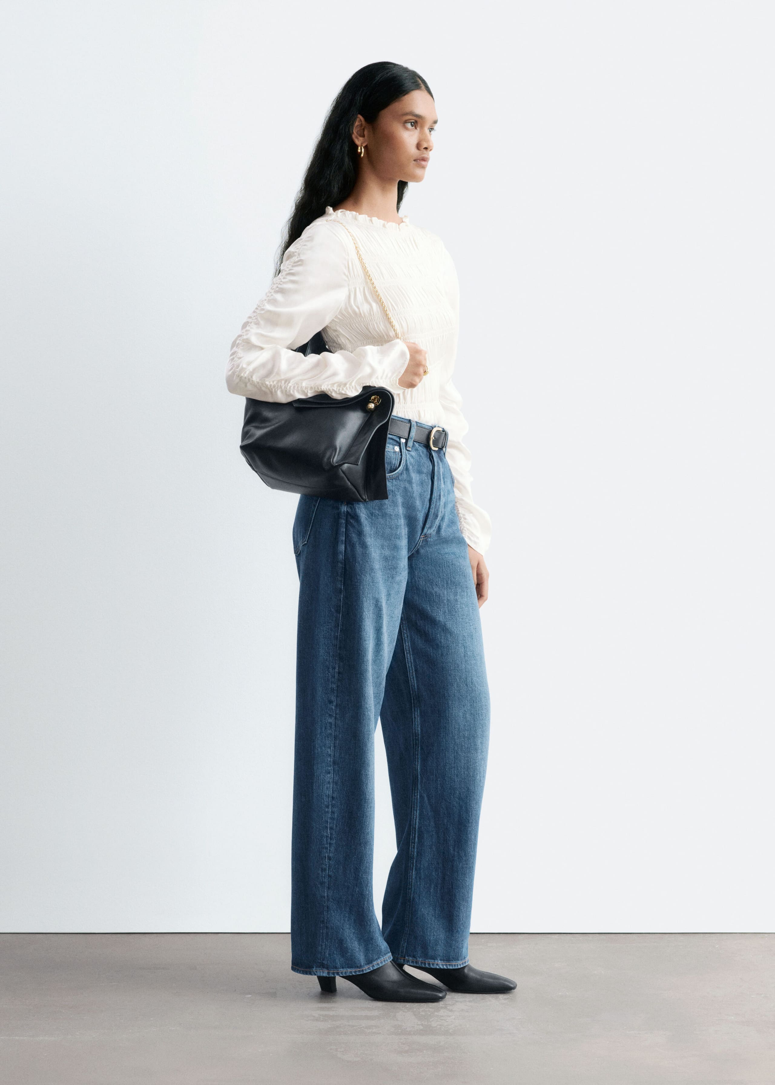 Blue denim jeans with a mid-rise waist and wide, barrel-shaped legs are worn by the model, who is standing in profile. The jeans are styled with a black leather belt, a tucked-in textured white blouse, and black pointed-toe boots.