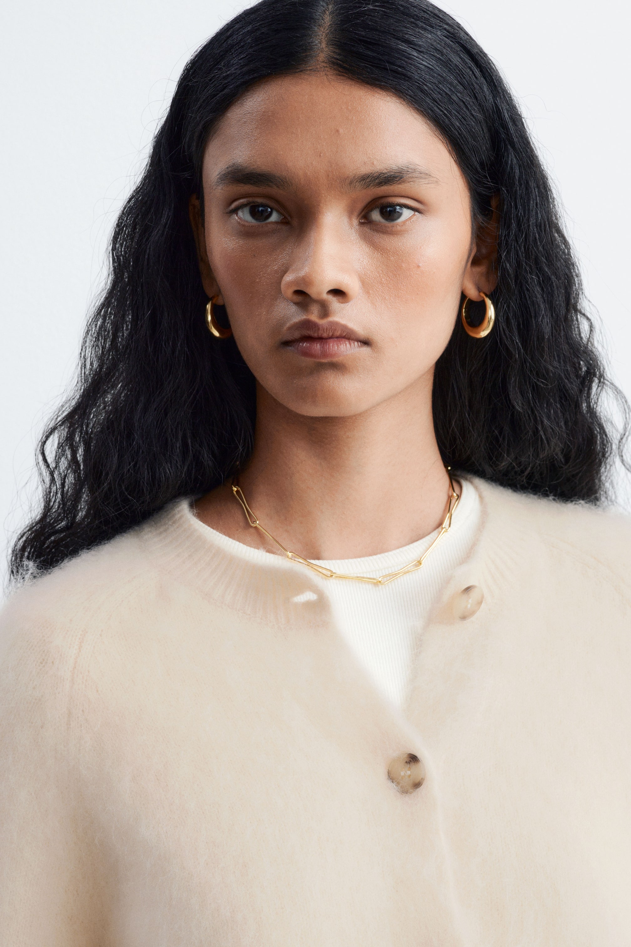 View larger image: A cream-coloured mohair-blend cardigan and a polished brass chain necklace with organic-shaped links are being worn by the model. The soft, fluffy cardigan is styled partially unbuttoned over a white top, highlighting the delicate gold-toned necklace.