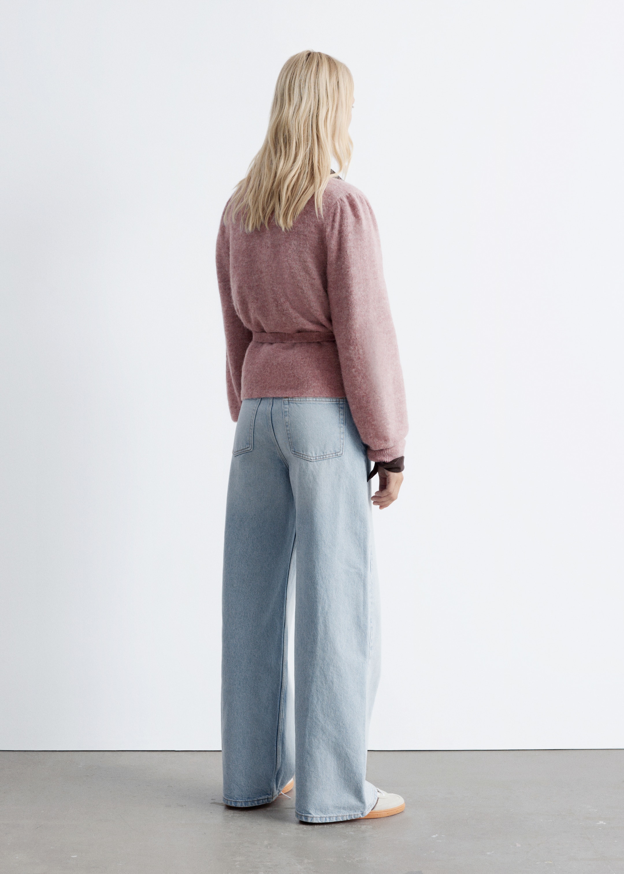 Ver imagem maior: Um casaco de malha rosa empoeirado com um design cruzado, cintura com laço e mangas compridas bufantes está a ser usado por uma modelo. A modelo está de costas para o espectador, a combinar o casaco com jeans de perna larga e lavagem clara.