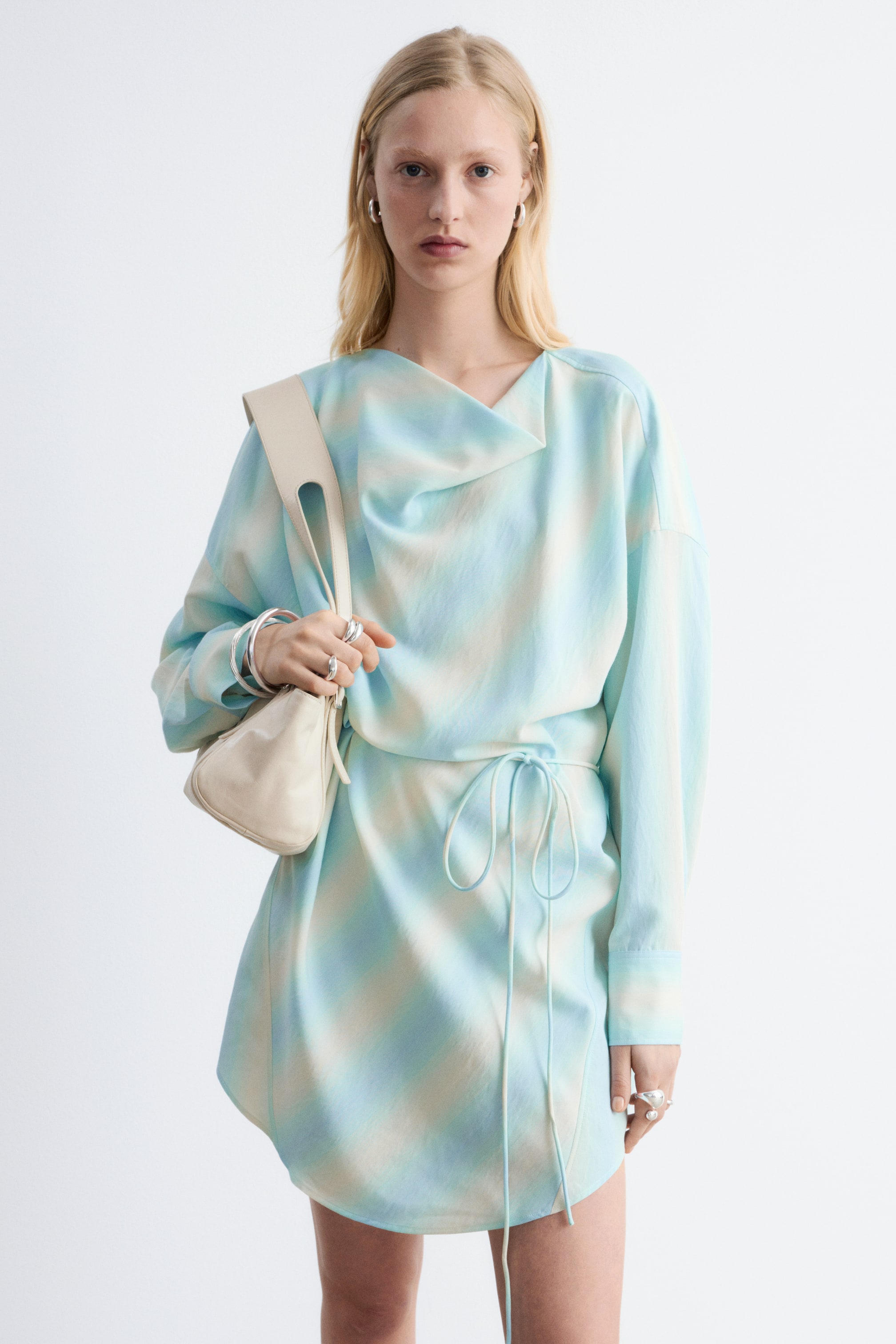 Größeres Bild ansehen: Ein locker sitzendes Minikleid zeigt ein abstraktes Muster in Hellblau und Creme, mit einem drapierten Ausschnitt, langen Ärmeln und einem asymmetrischen Saum. Es ist mit einer Bindegürtel und sichtbaren Kordeln versehen.