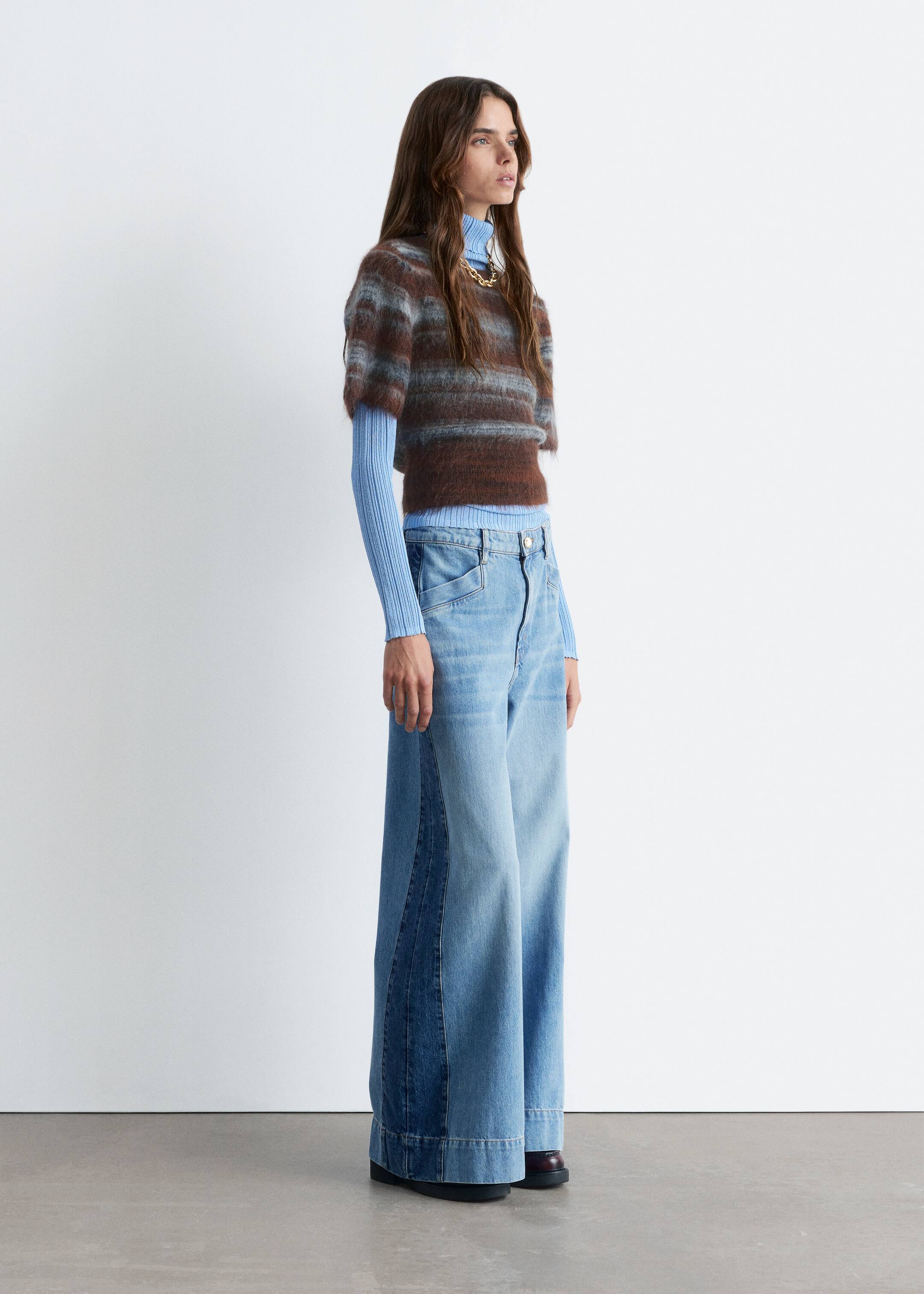 Agrandir l'image: Une tenue est présentée de côté, mettant en avant un pull court à manches courtes et duveteux avec des rayures horizontales marron et bleu clair, superposé à un col roulé côtelé bleu clair. Un collier à maillons épais de ton doré orne le col roulé, tandis qu'un jean à taille haute, à jambes larges et délavé clair avec des panneaux de denim plus foncé le long des coutures extérieures est porté, cachant partiellement des chaussures à talons bas et foncées.