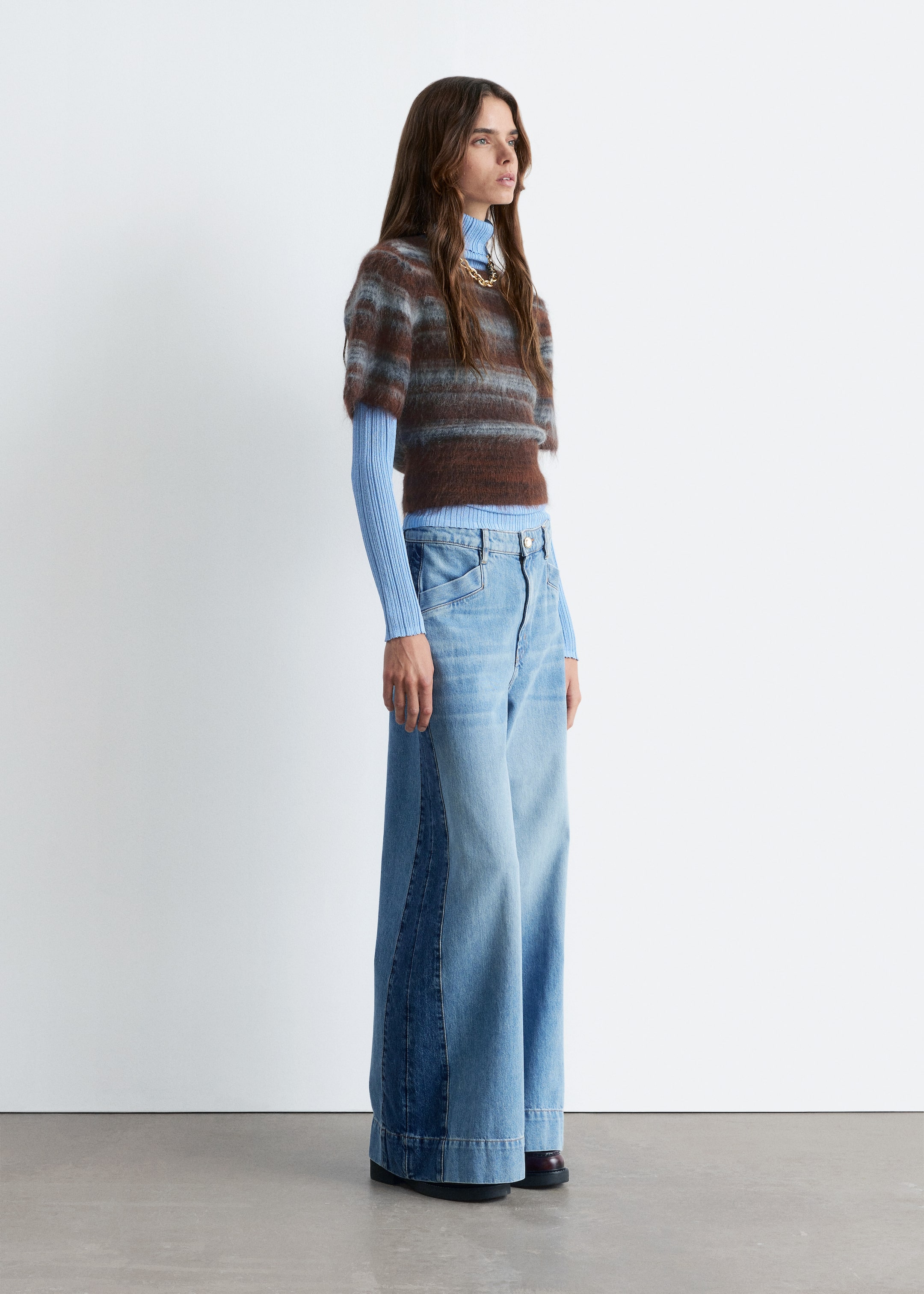 Größeres Bild ansehen: Ein Outfit wird von der Seite präsentiert, mit einem kurzärmeligen, flauschigen Pullover mit horizontalen braunen und hellblauen Streifen, der über einem hellblauen gerippten Rollkragenpullover getragen wird. Eine goldfarbene, klobige Kettenhalskette schmückt den Rollkragen, während hell gewaschene, hoch taillierte, weit geschnittene Jeans mit dunkleren Denim-Paneelen entlang der äußeren Nähte getragen werden, die teilweise dunkle Schuhe mit niedrigem Absatz verdecken.