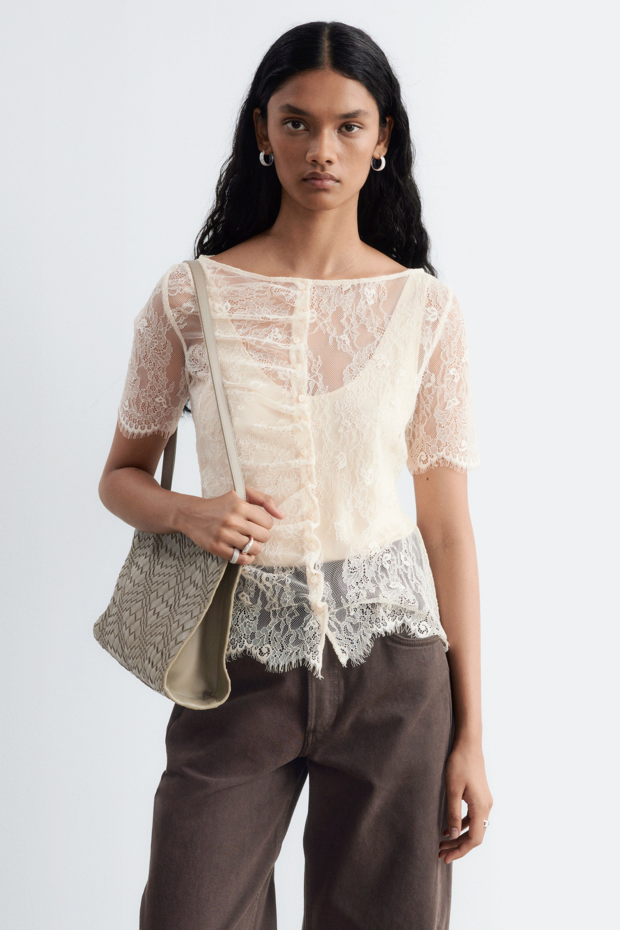 Grotere afbeelding bekijken: Een crèmekleurige top van kant met een slank silhouet, boothals, korte mouwen met geschulpte randen en een geknoopte voorkant, wordt door het model over een camisole gedragen.