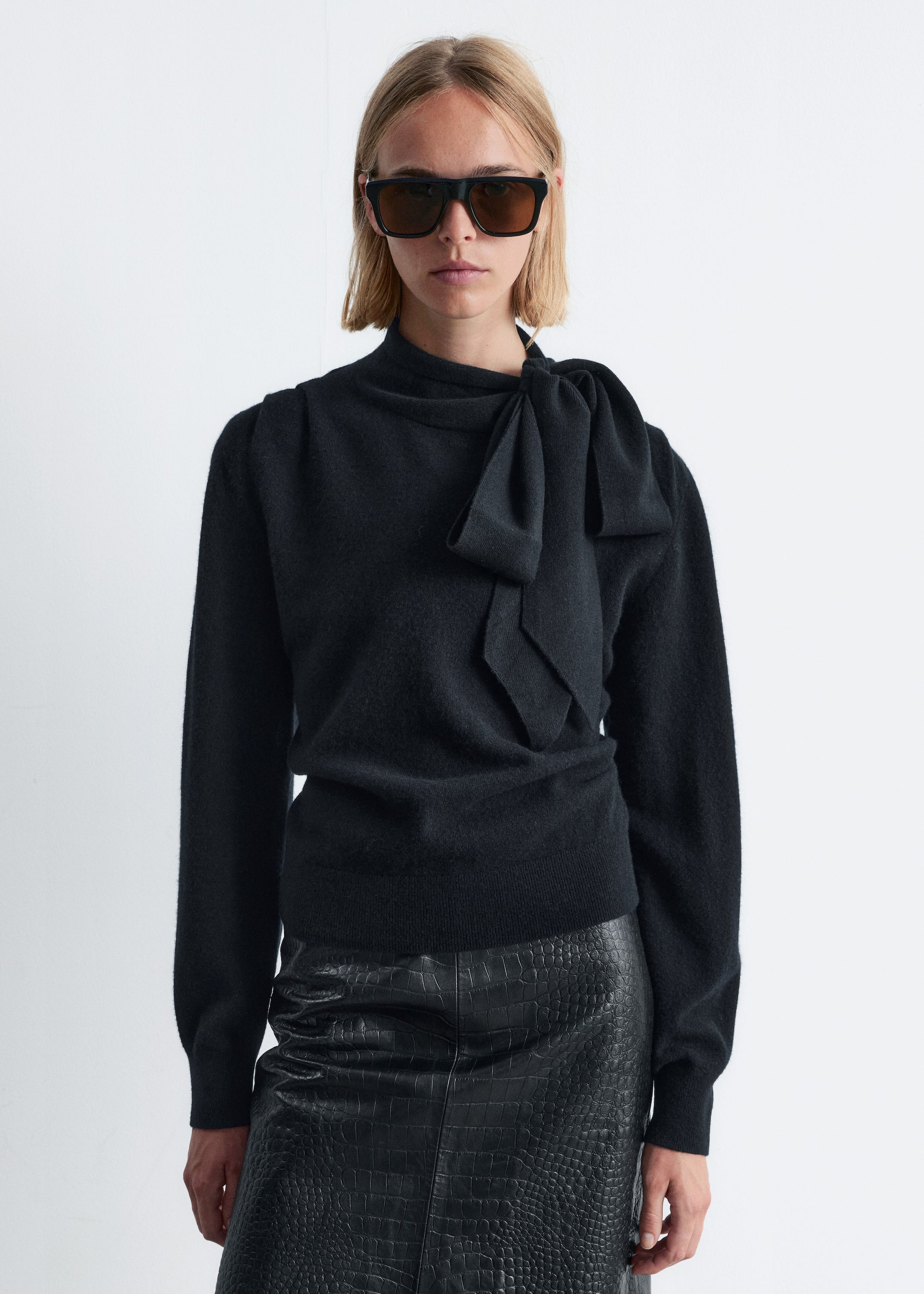 Größeres Bild ansehen: Ein schwarzer Strickpullover wird getragen, der einen hohen Kragen und ein großes, integriertes Schleifendetail aufweist, das auf der linken Schulter gebunden ist. Er präsentiert lange Ärmel mit subtilen Raffungen an den Schultern und eng anliegenden, gerippten Bündchen, wobei sein gerippter Saum auf der Hüfte sitzt.