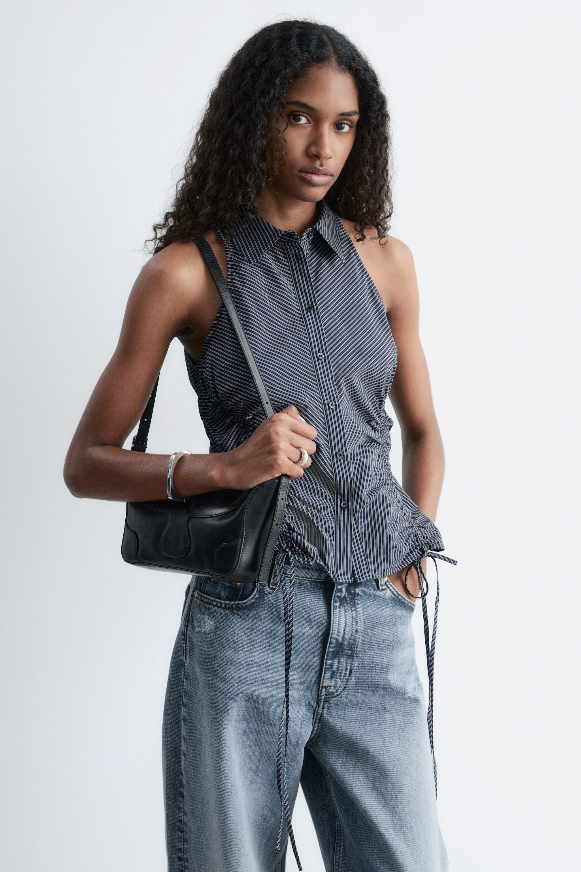 View larger image: A sleeveless collared top featuring a dark and light striped pattern, with diagonal stripes on the bodice transitioning to vertical stripes on the lower front. It is designed with a button-through closure, a pointed collar, and adjustable drawstrings at the side seams that create a ruched effect and fall over light-wash jeans.